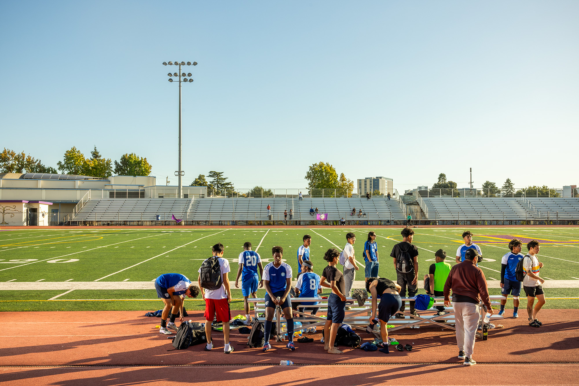 Fotbalisté v modrých dresech a kopačkách mluví se svým trenérem za slunečného dne na fotbalovém hřišti.