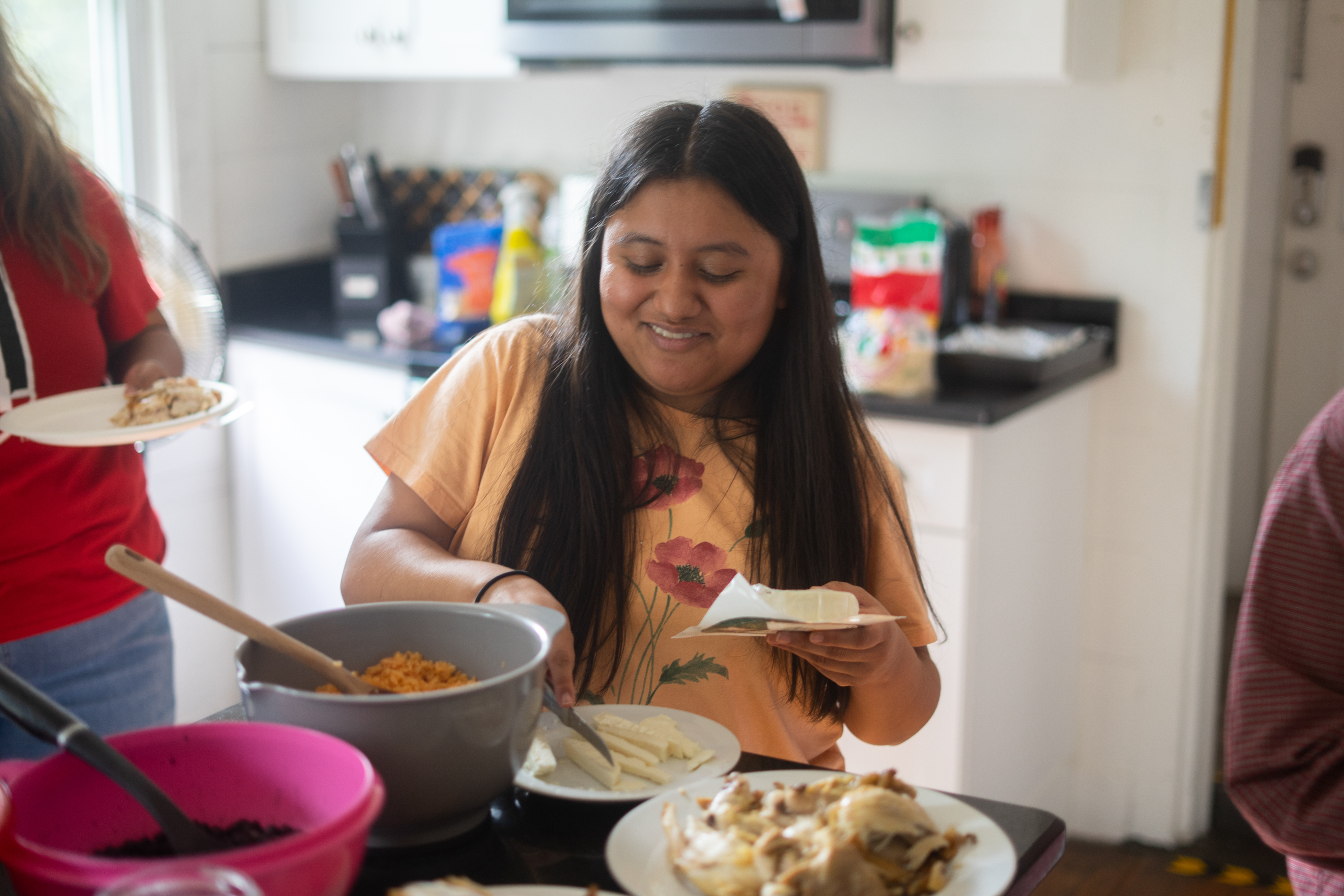 A woman with medium-toned skin and long dark hair, wearing a peach-colored top with flowers on it, is seated in a kitchen slicing cheese. A plate of chicken and a bowl of rice are on the counter in front of her, and a woman in a red T-shirt stands to her right.