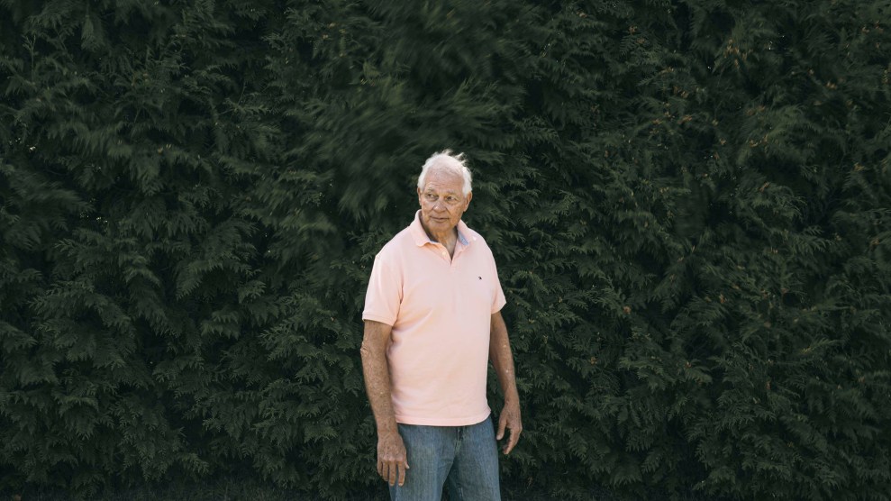 A white man wearing a pink shirt standing in front of a green hedge.