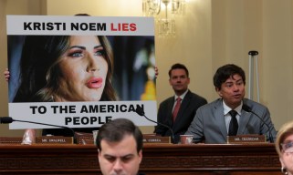 Shri Thanedar, a South Asian man with black hair in a gray suit and black tie, sites next to a poster with an image of Kristi Noem and the text "Kristi Noem Lies to the American People."