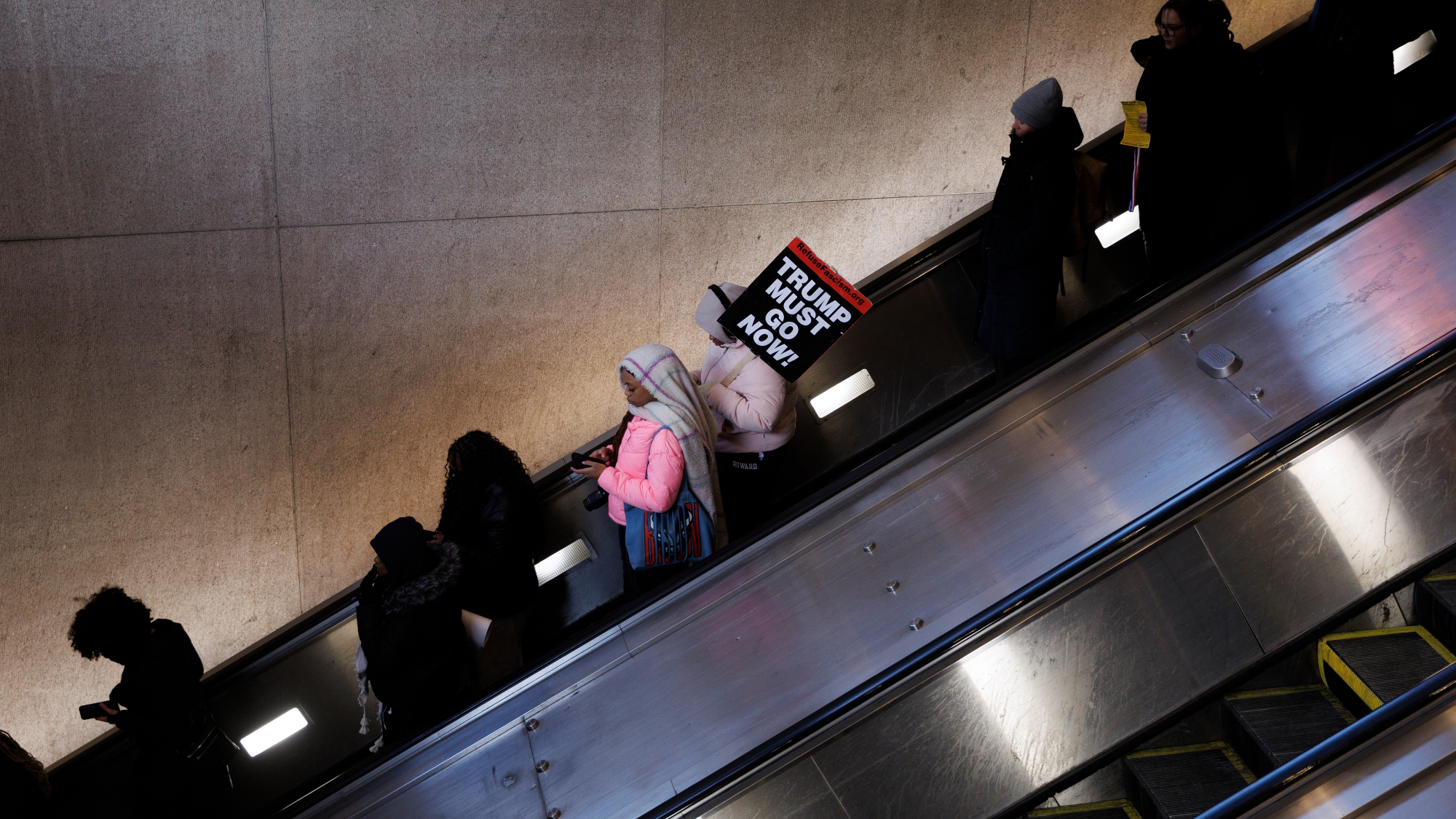 emonstrators descend on an escalator after attending a protest. A sign reads "TRUMP MUST GO NOW"
