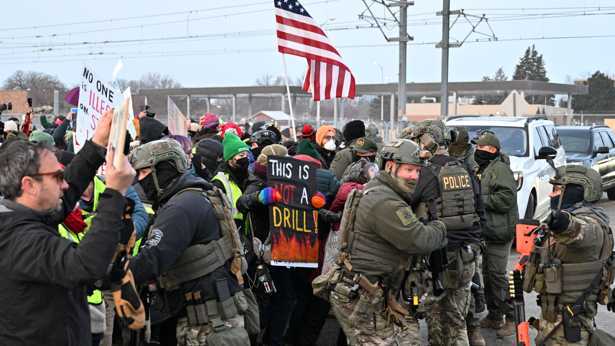A group of protesters gathered holding signs, with police in riot gear pushing them back.
