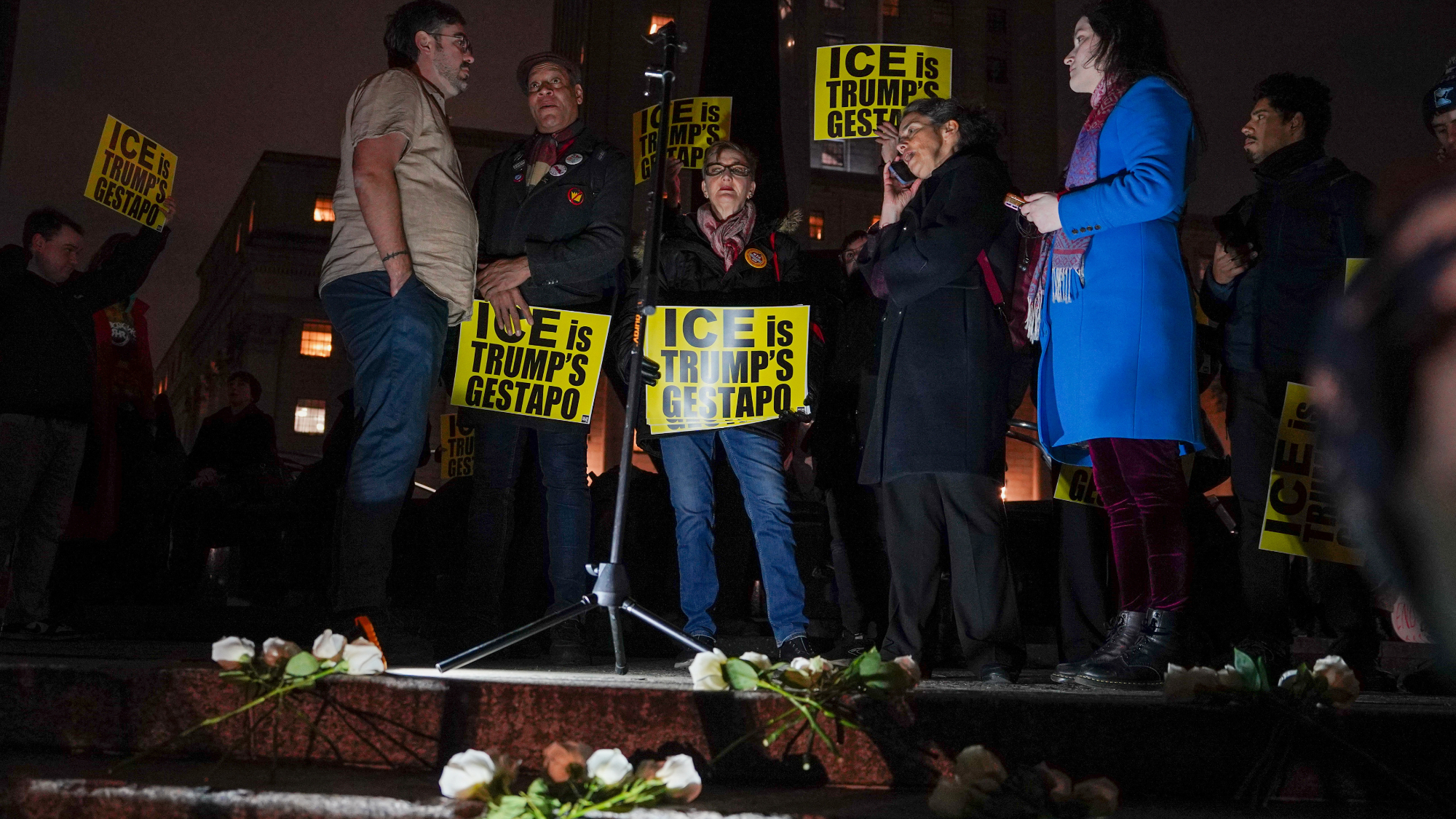 People gathered in the dark, holding yellow signs that say "ICE is Trumo's Gestapo"