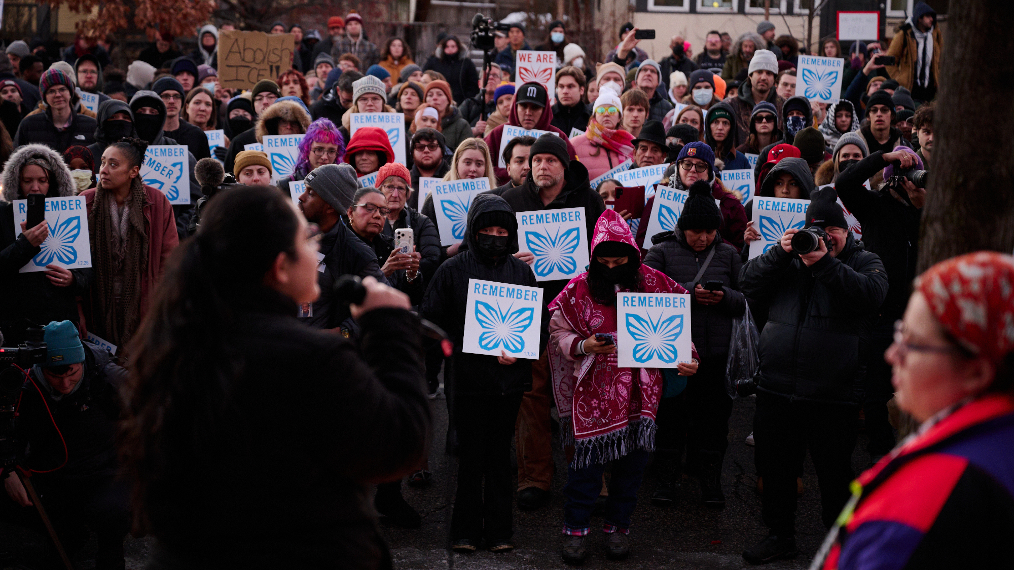 Community members gather for an emergency vigil, holding up signs with a butterfly that says "remember."