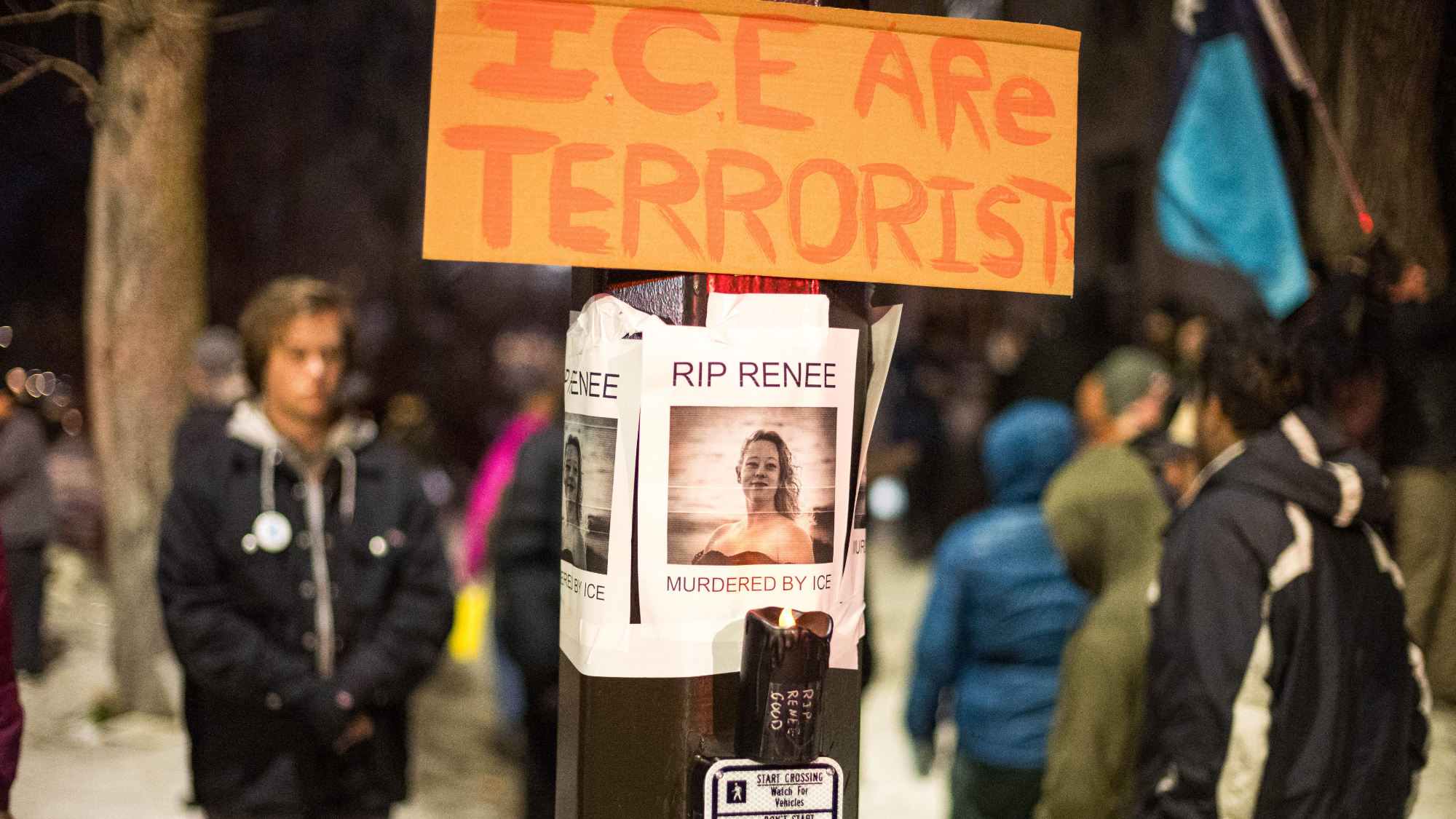 Signs that say "ICE Are Terroris" and "RIP Renee" on a pole outside with people in the background.