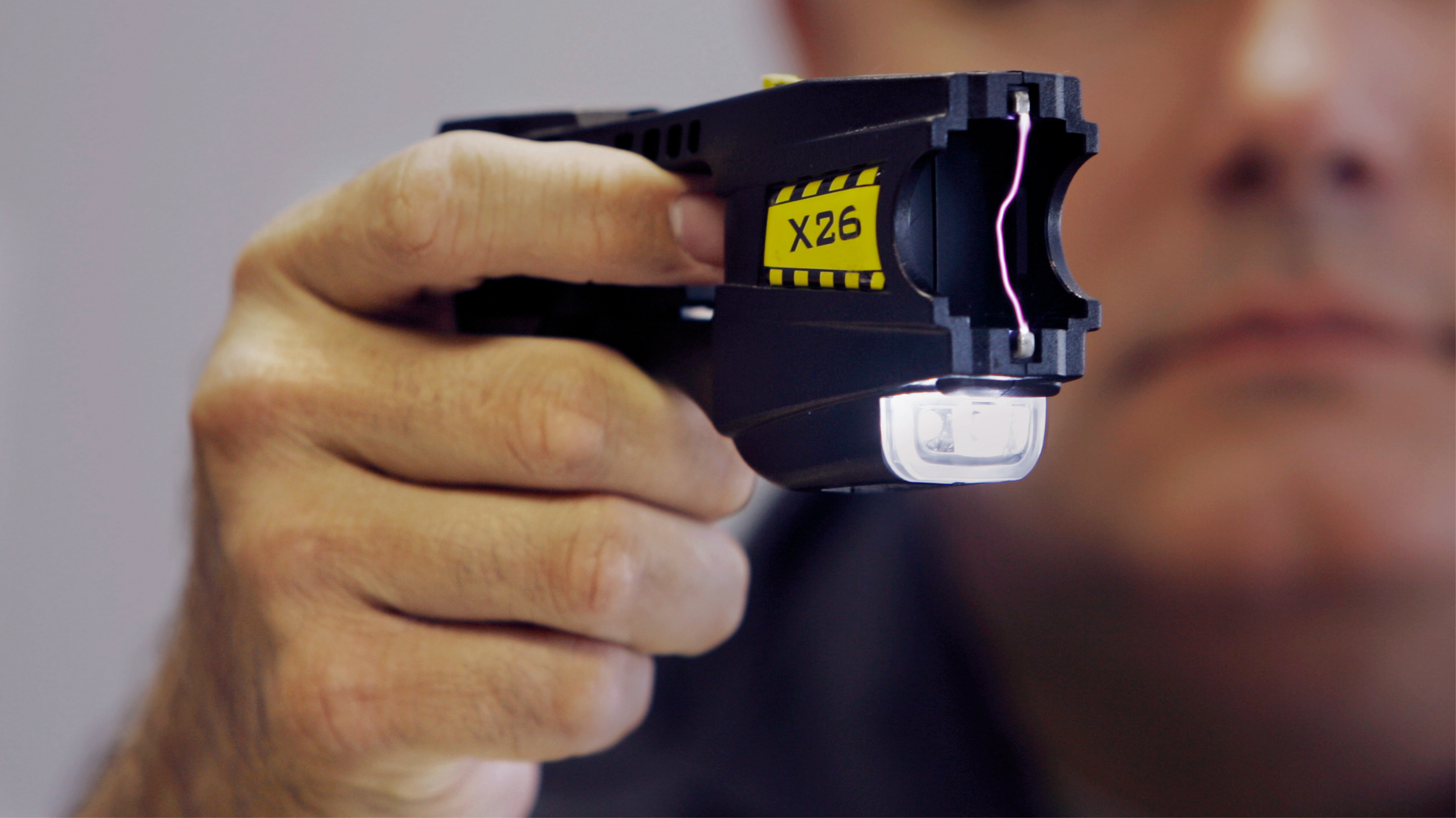 A man’s hand squeezes a black stun gun, which is emitting a thin electrical arc at the tip.