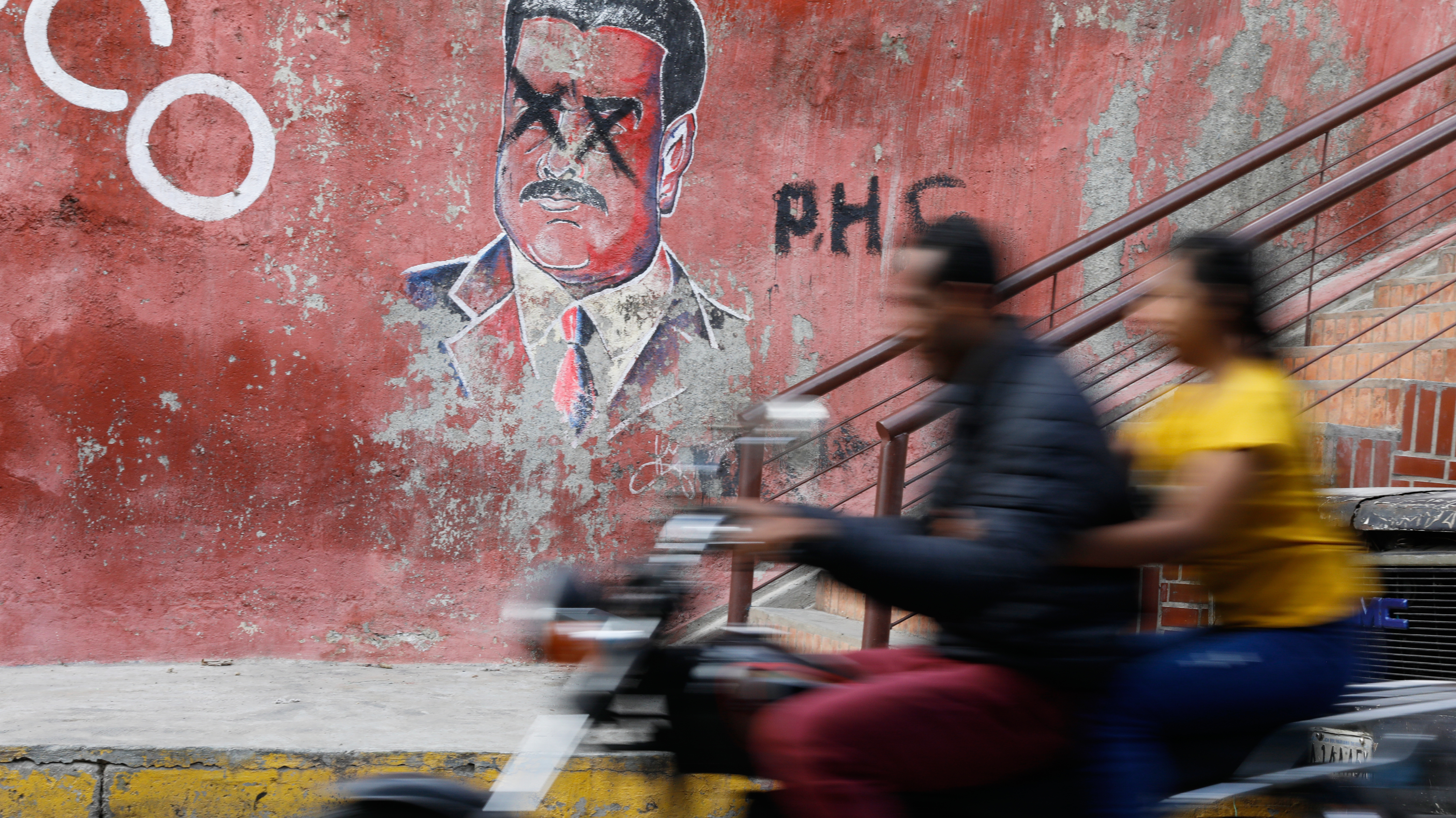 A man and woman on a motorcycle are blurry in the foreground as they ride past a mural of Nicolás Maduro on a red concrete wall. Two black Xs are spray-painted over the eyes of Maduro’s portrait.
