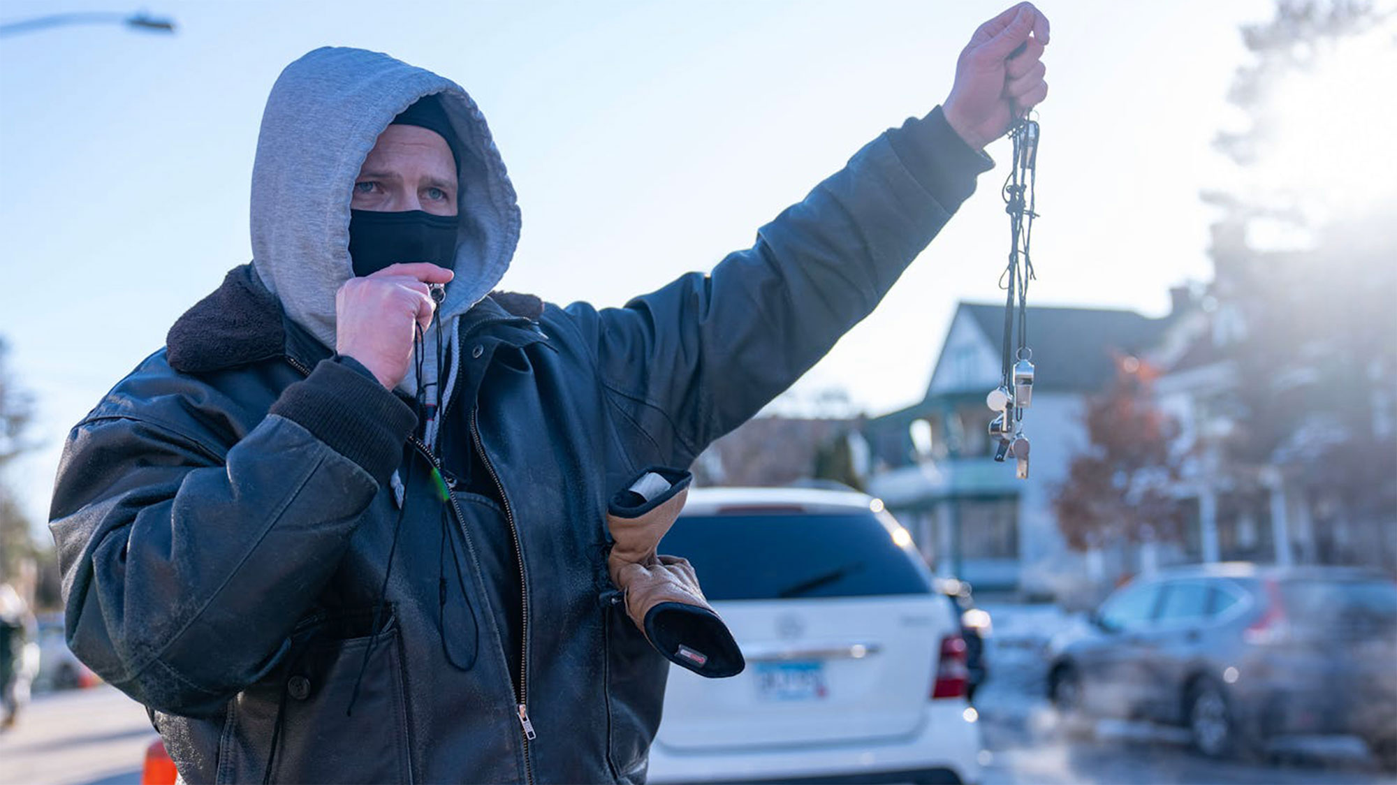 On a sunny day in Minneapolis, a white man, face partially masked, in a dark jacket over a light gray hoodie, holds out a handful of whistles dangling from strings.