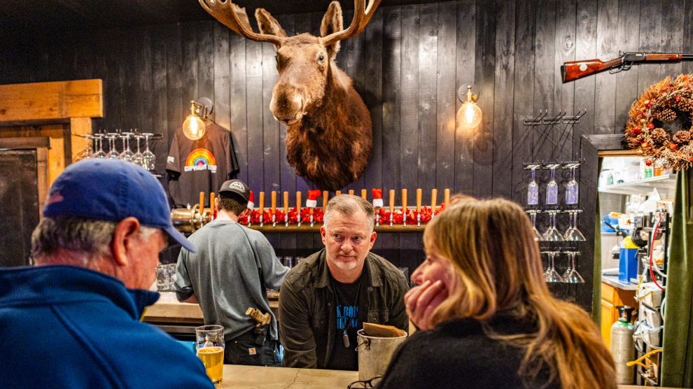 Four people dressed in warm, long-sleeved clothing at a wood-paneled bar with a moose head and rifle mounted on the wall. Behind the bar, a man with a gun on his hip stands facing a row of beer taps, and another looks attentively at a man and woman seated at the bar.