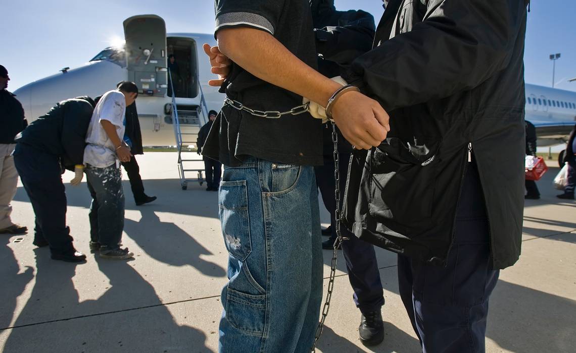 Close-crop of men being shackled around the wrist and waist by an officer on the tarmac outside of a plane. 