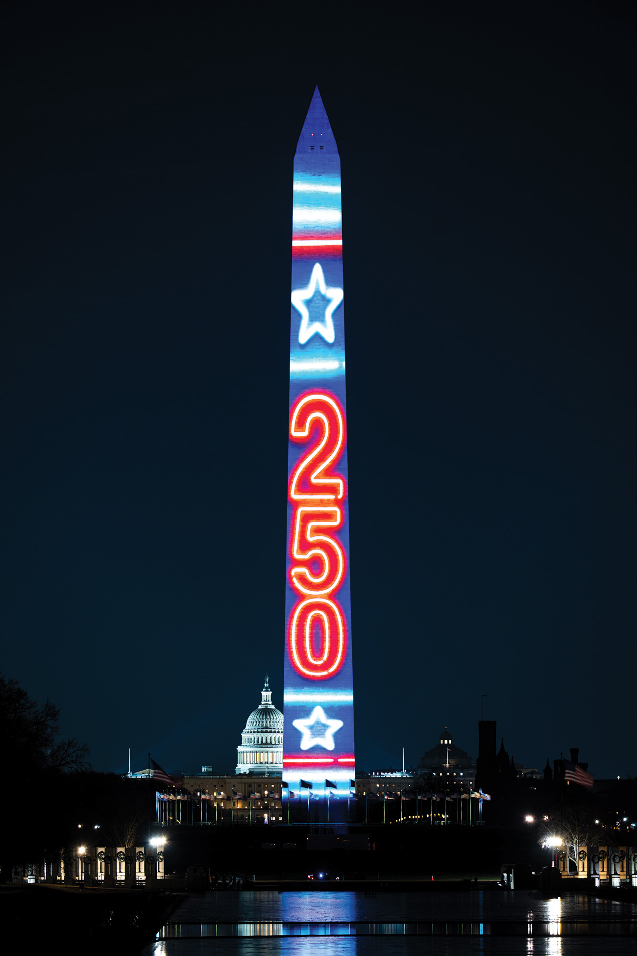 A night view of the Washington Monument illuminated with a light projection featuring horizontal blue bands, white stars, and the red numbers "250" arranged vertically. The lit dome of the U.S. Capitol is visible in the background, with the reflecting pool in the foreground.