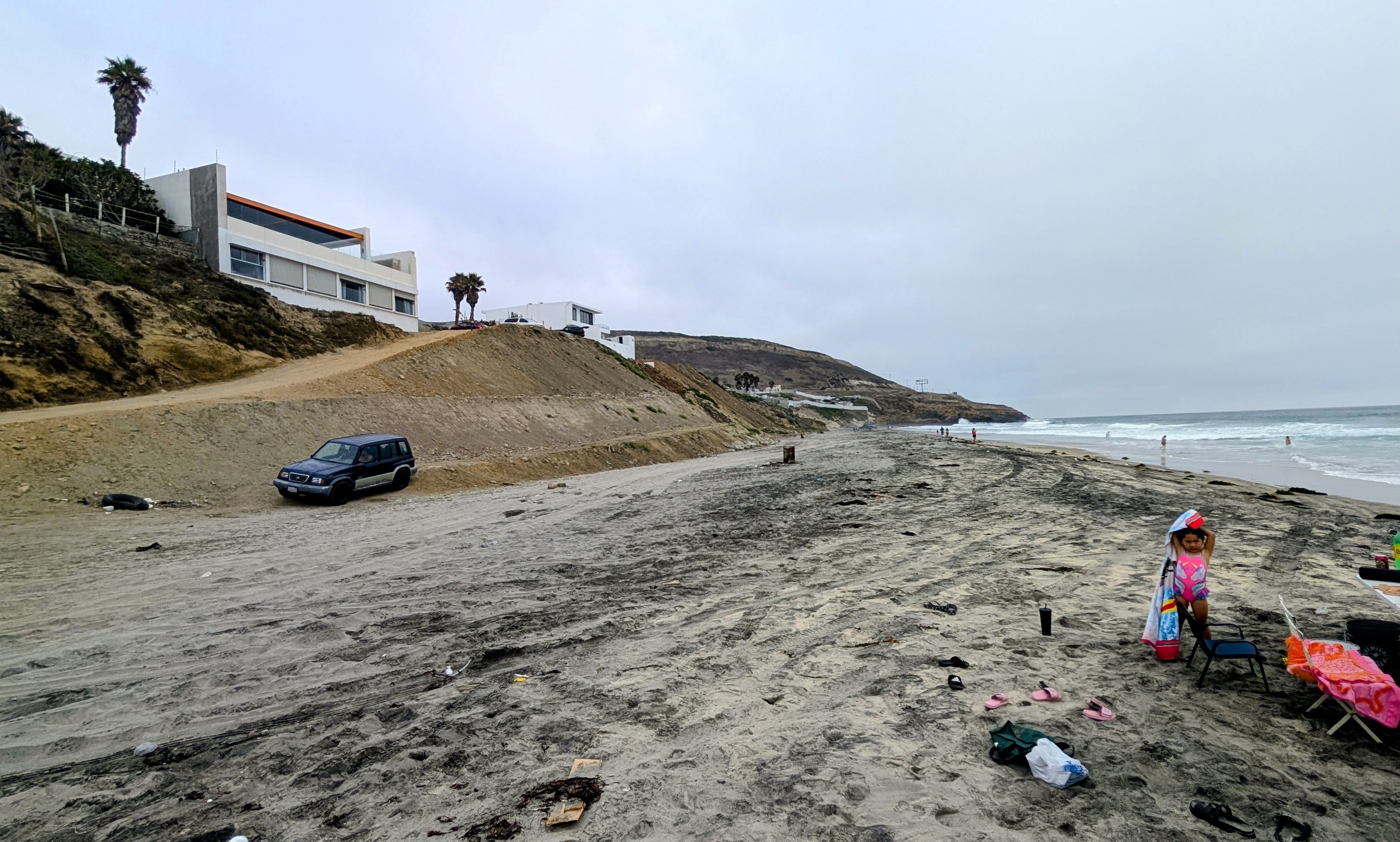 The beach on a cloudy day, with a handful of people in the water, in front of a few buildings.