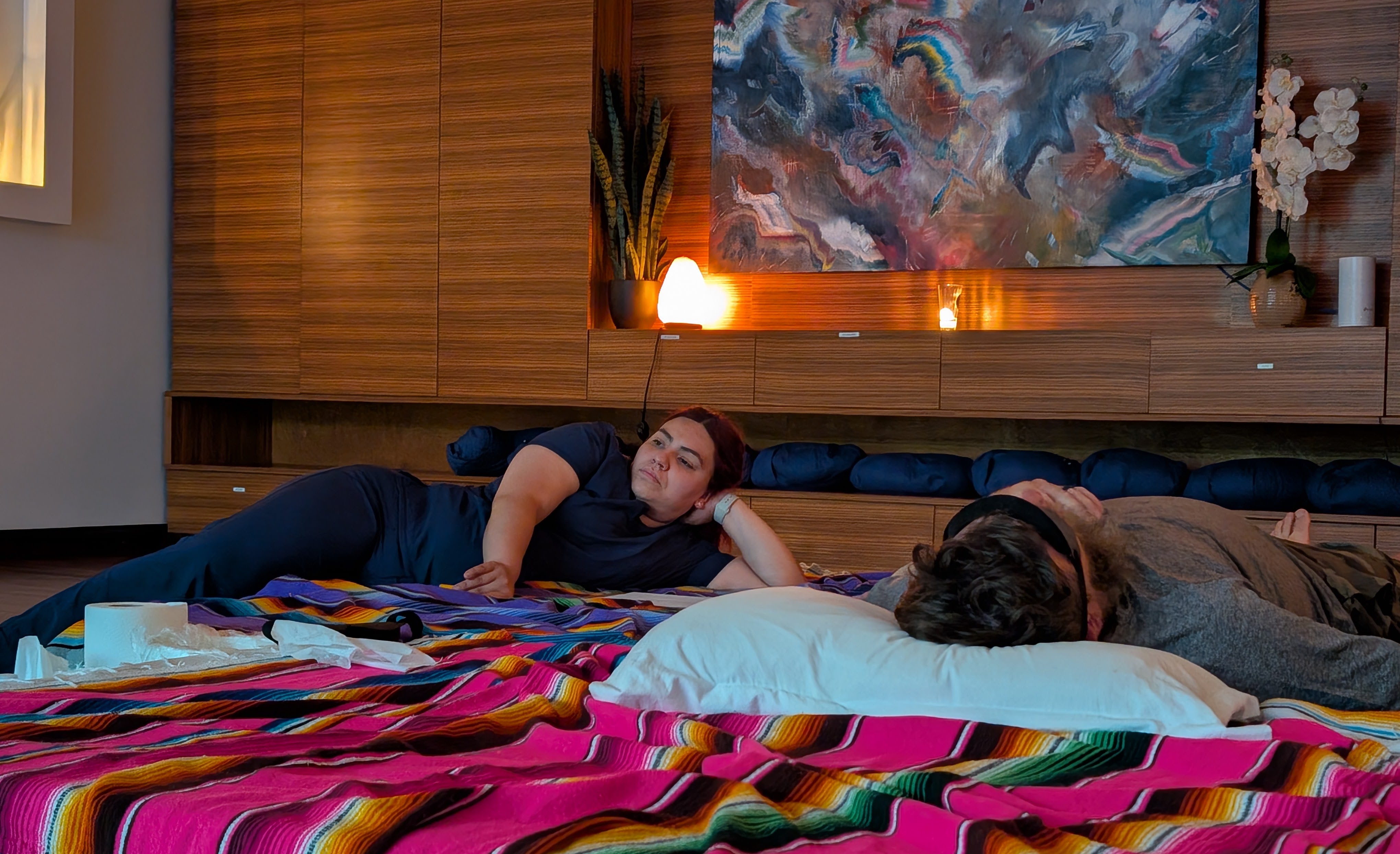 A nurse lays next to a man wearing an eye mask on a colorful blanket.