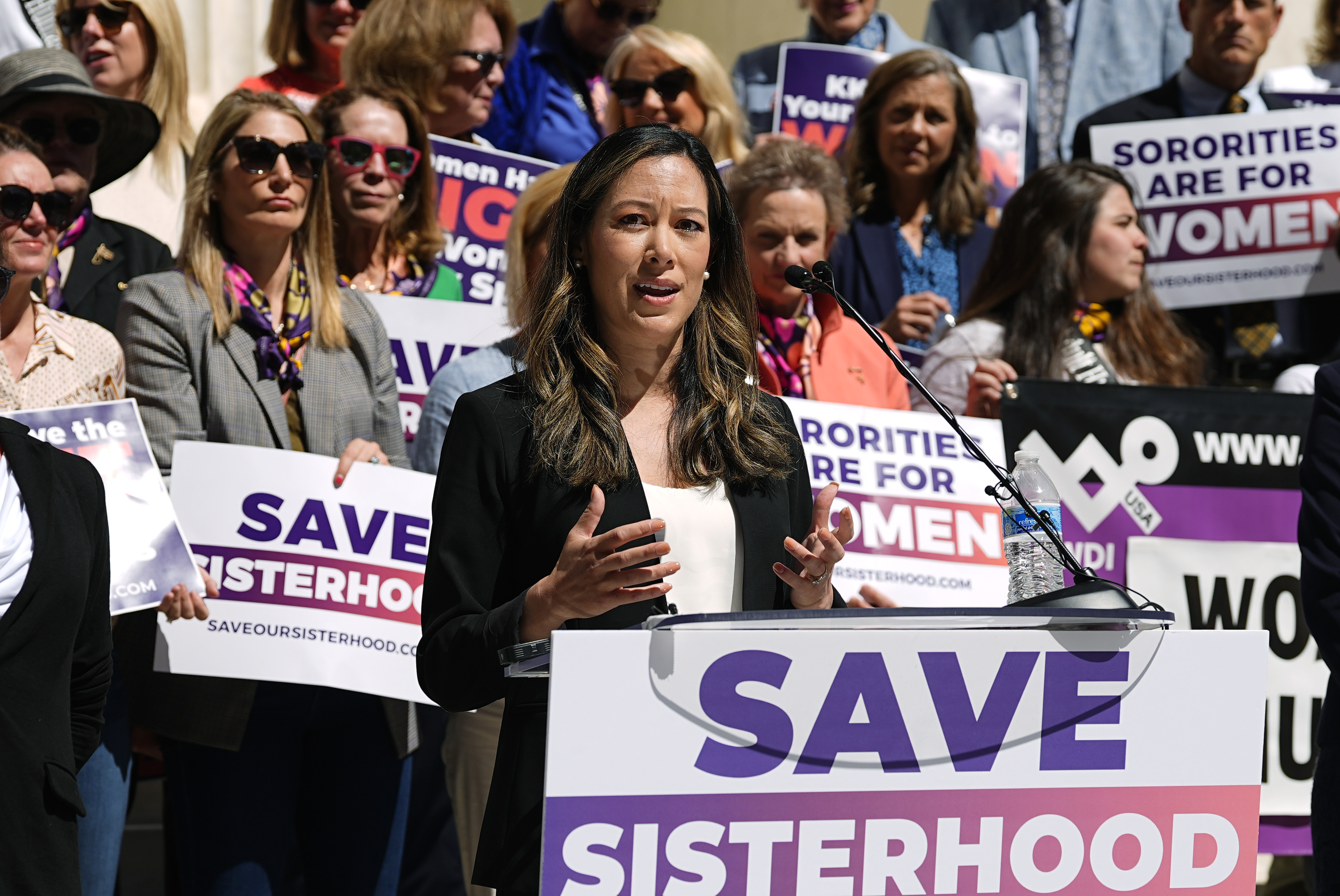 A young woman with long dark brown hair speaks on a podium with a sign that reads, "Save Sisterhood," outside a courthouse alongside protesters.