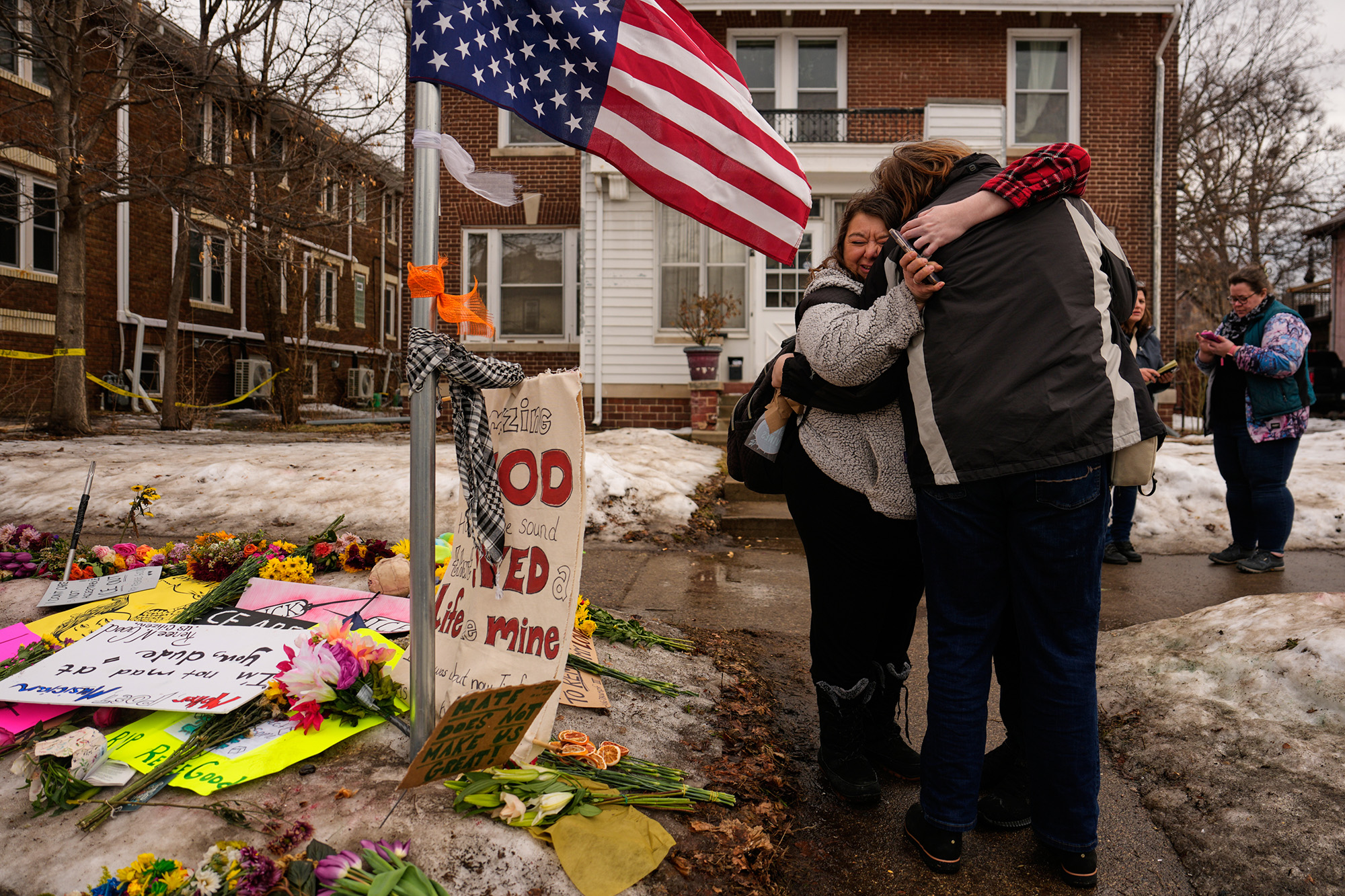 Two people hug under and American flag, next to a memorial for Renee Good.