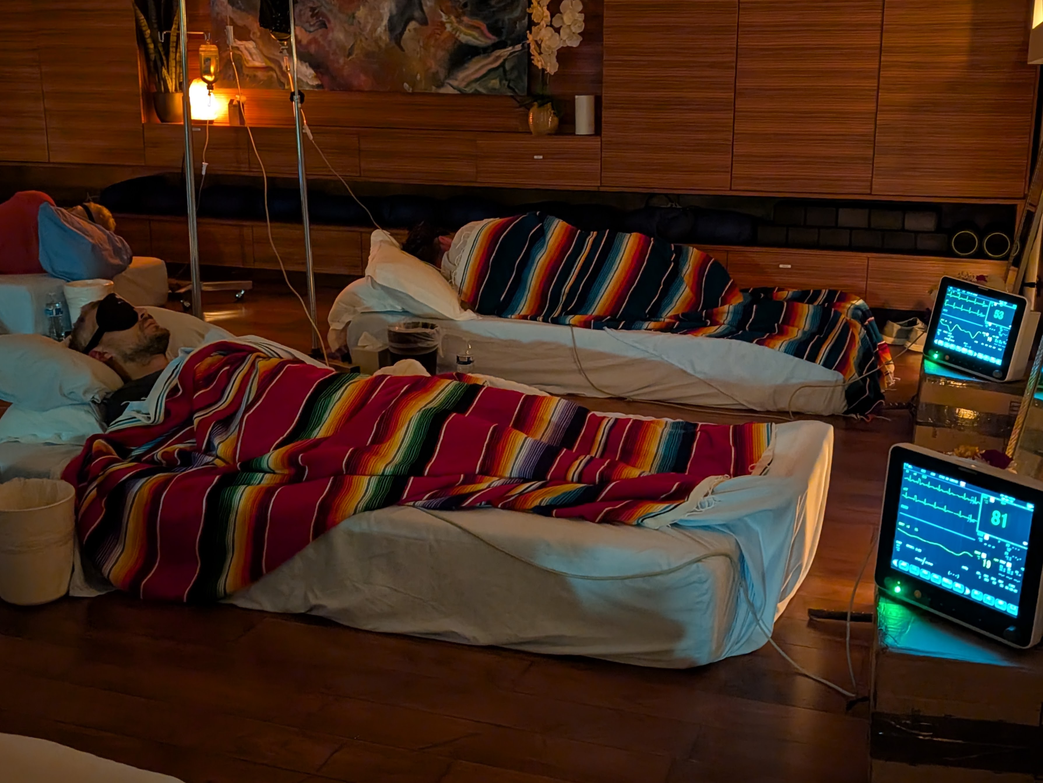 Three people lay on mattresses on the floor, covered in colorful blankets, are hooked up to EKGs and IVs.