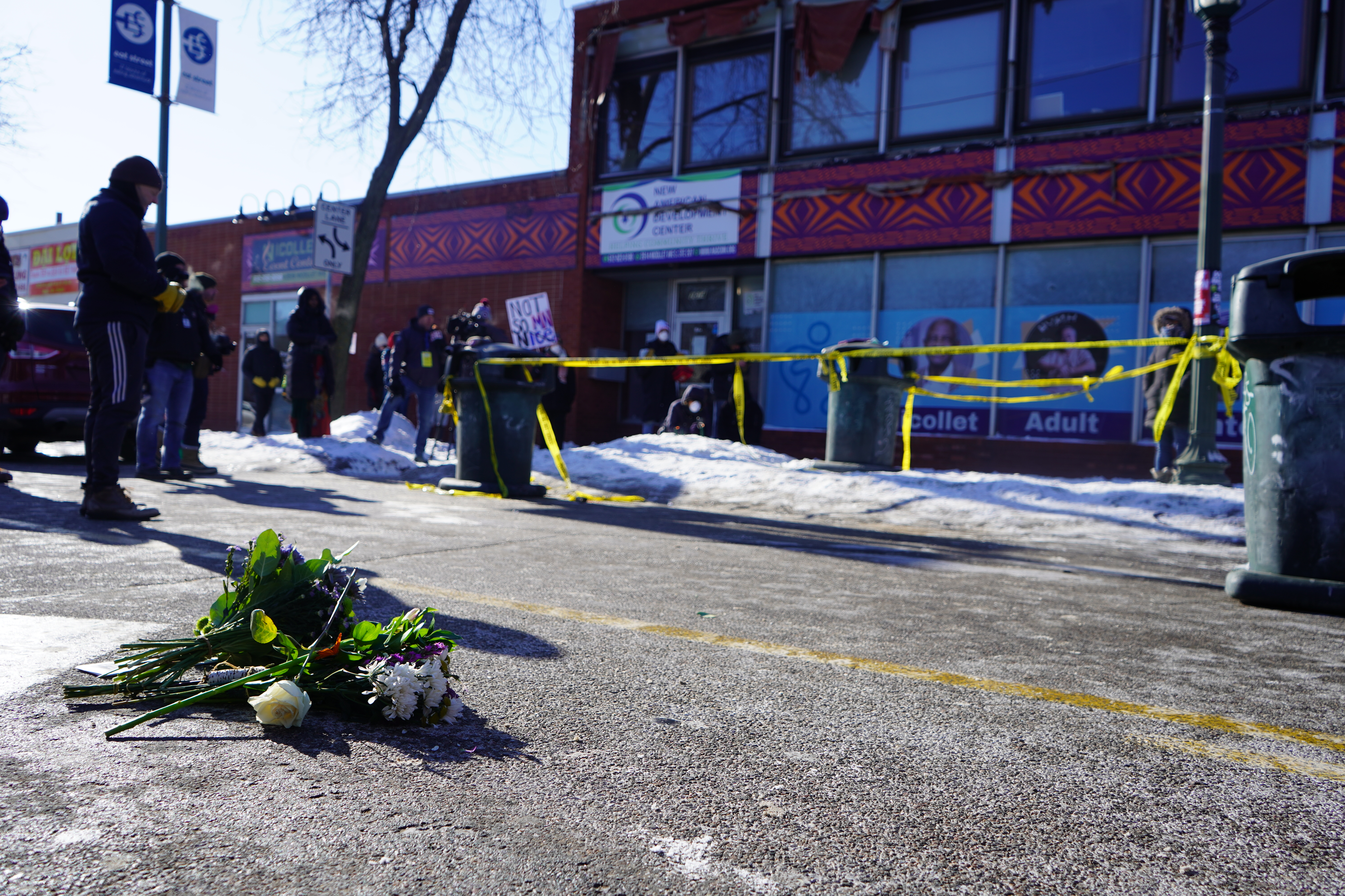 A small bouquet of flowers lies on a winter street in Minneapolis in the foreground, while yellow police tape cordons off the sidewalk and bundled-up community members gather near a commercial building in the background.