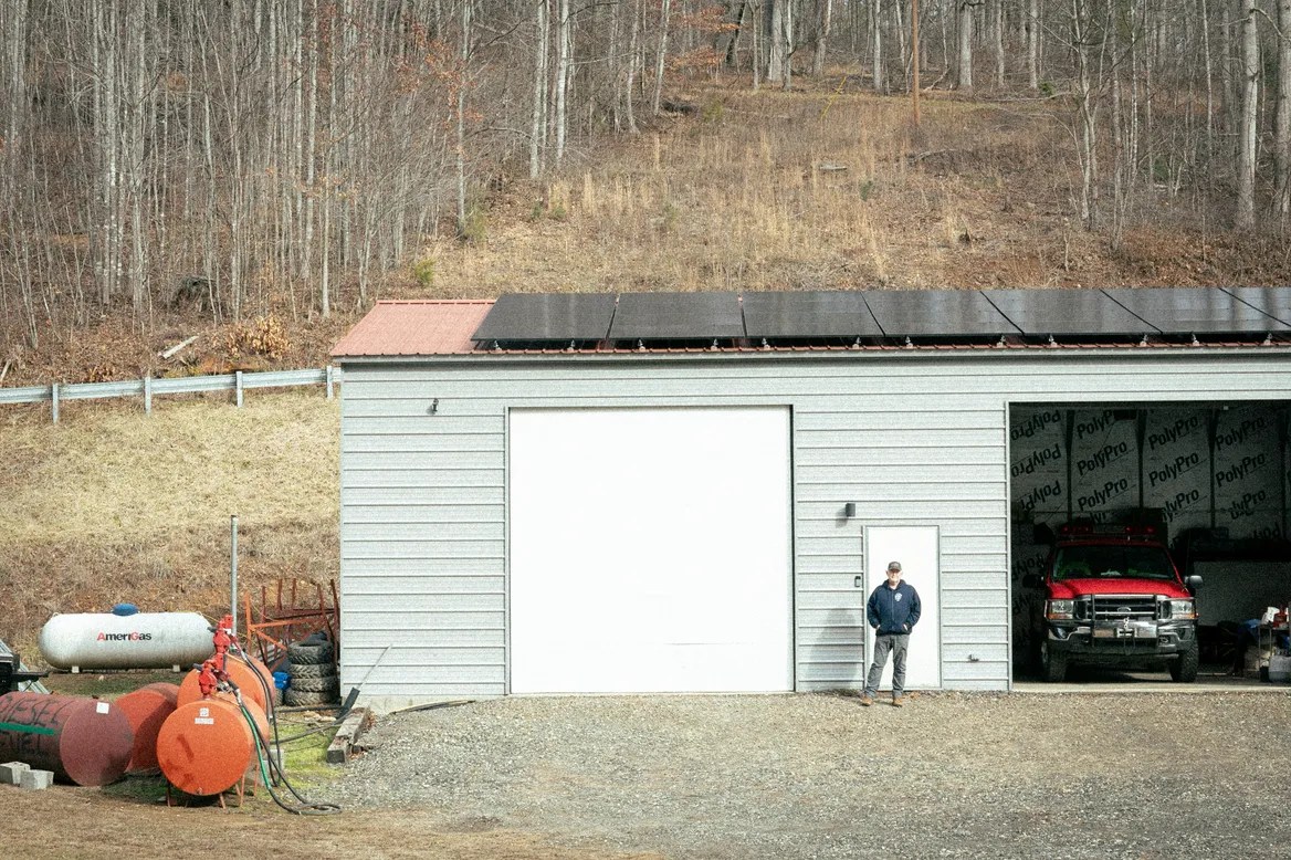 A man stands in front of a grey building adorned with solar panels.