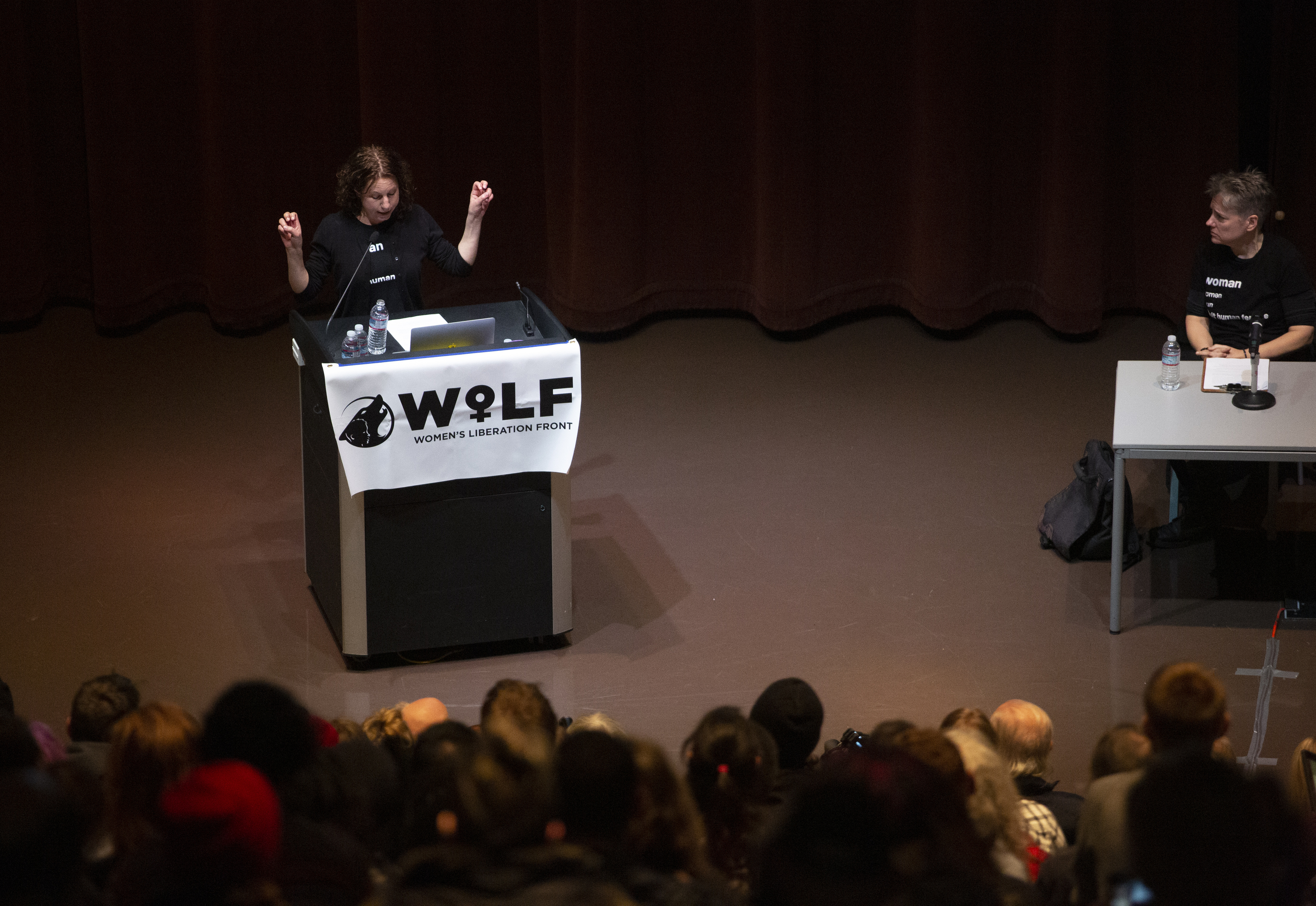A dark-haired woman speaks on a podium, making air-quote gestures with her hands.
