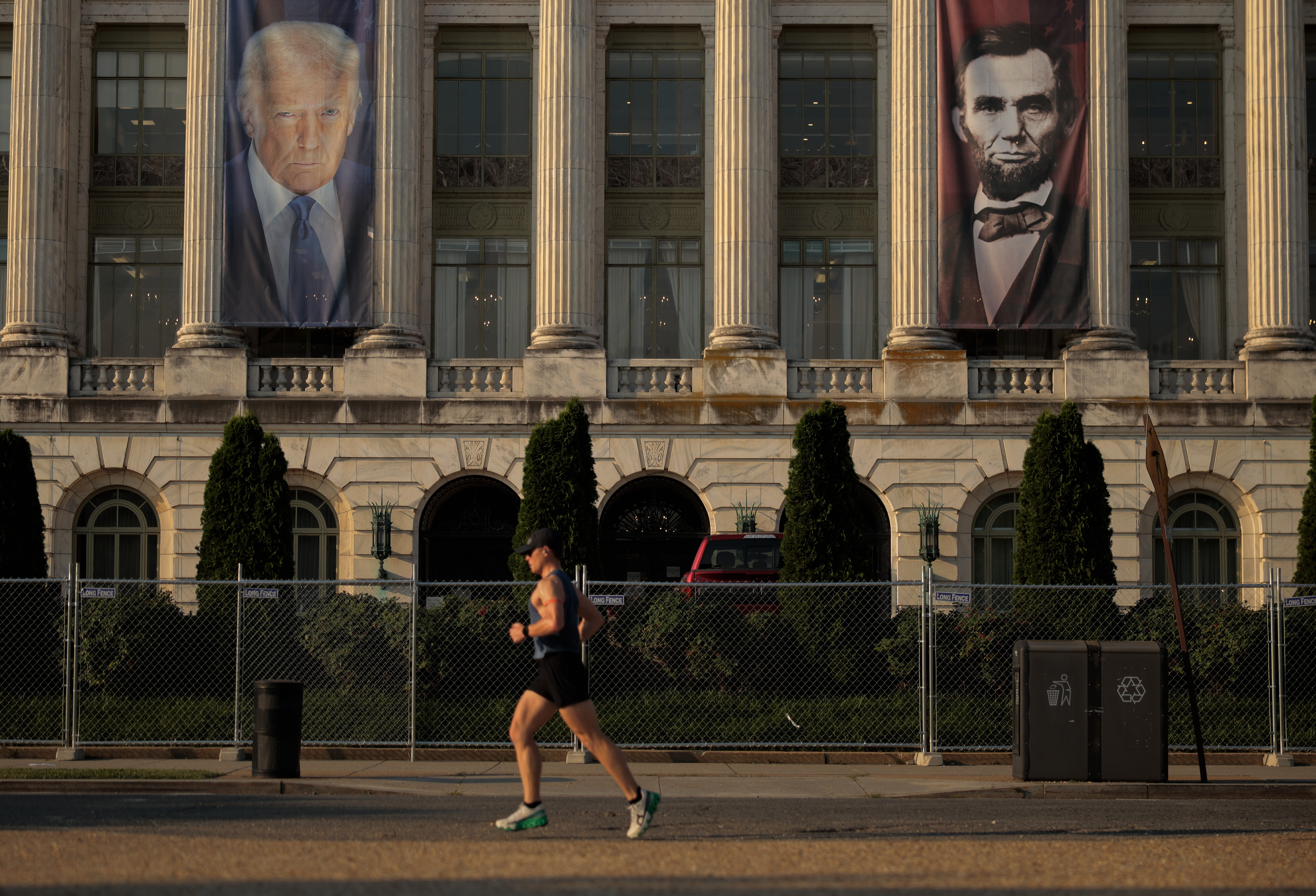 The facade of a neoclassical building with large columns, featuring two tall banners hanging between them: one of Donald Trump on the left and one of Abraham Lincoln on the right. In the foreground, a person in athletic gear is jogging along the street past a chain-link fence and green shrubbery.