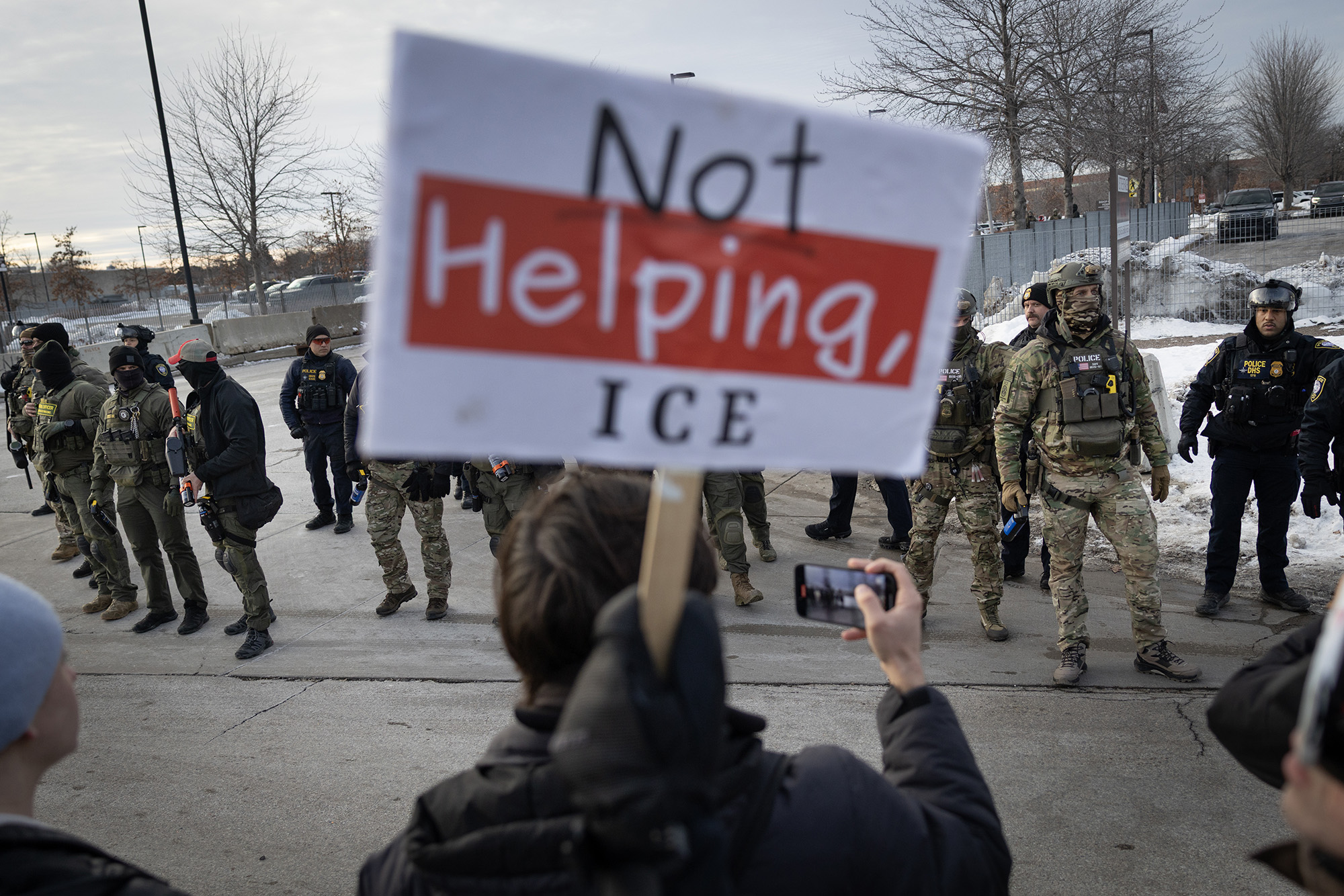 Person holding a sign that reads, "Not Helping, ICE" in front of a line of federal agents.