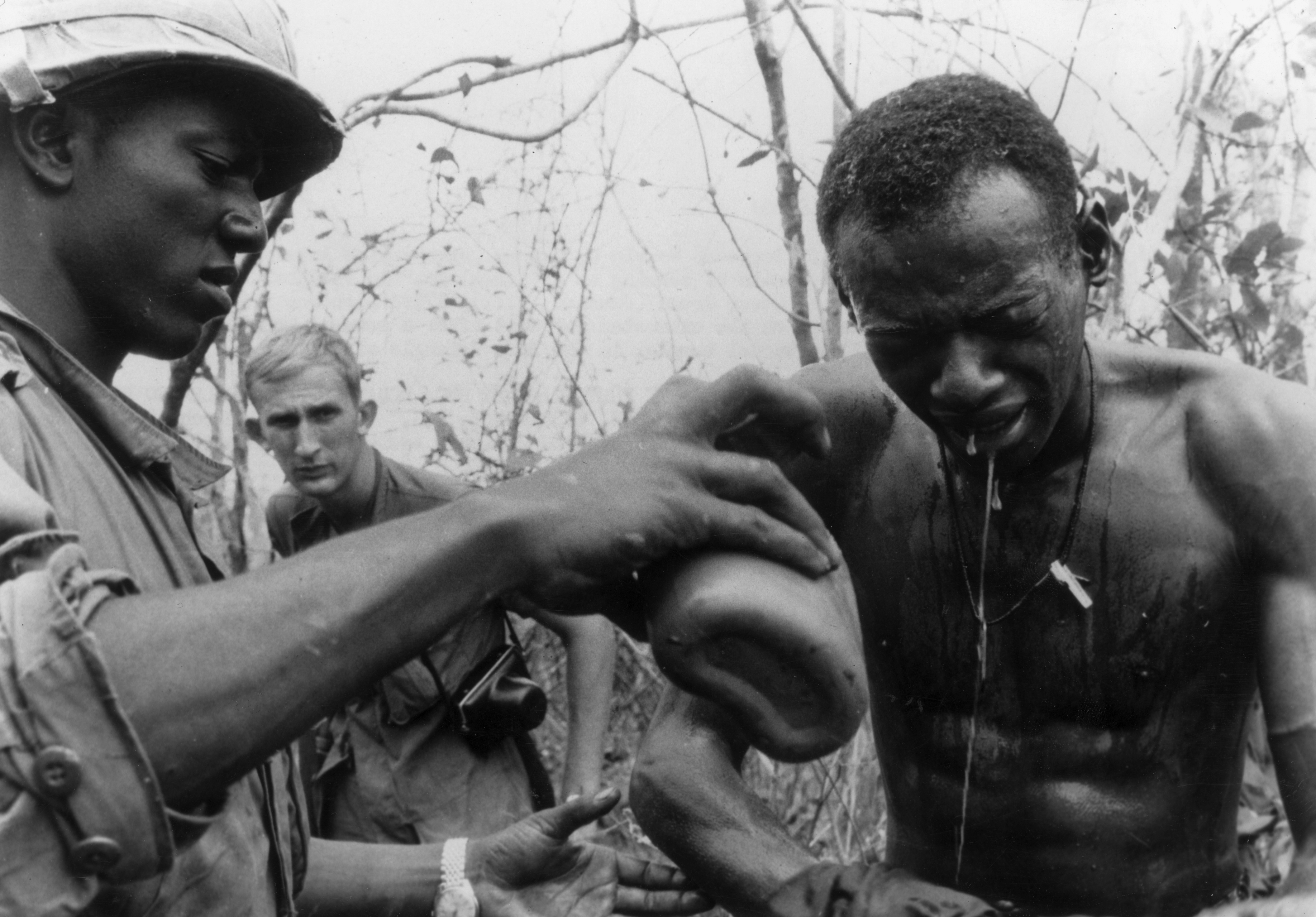 A Black soldier pours a canteen of water over another Black soldier's face in Vietnam. A white soldier looks on from the background.