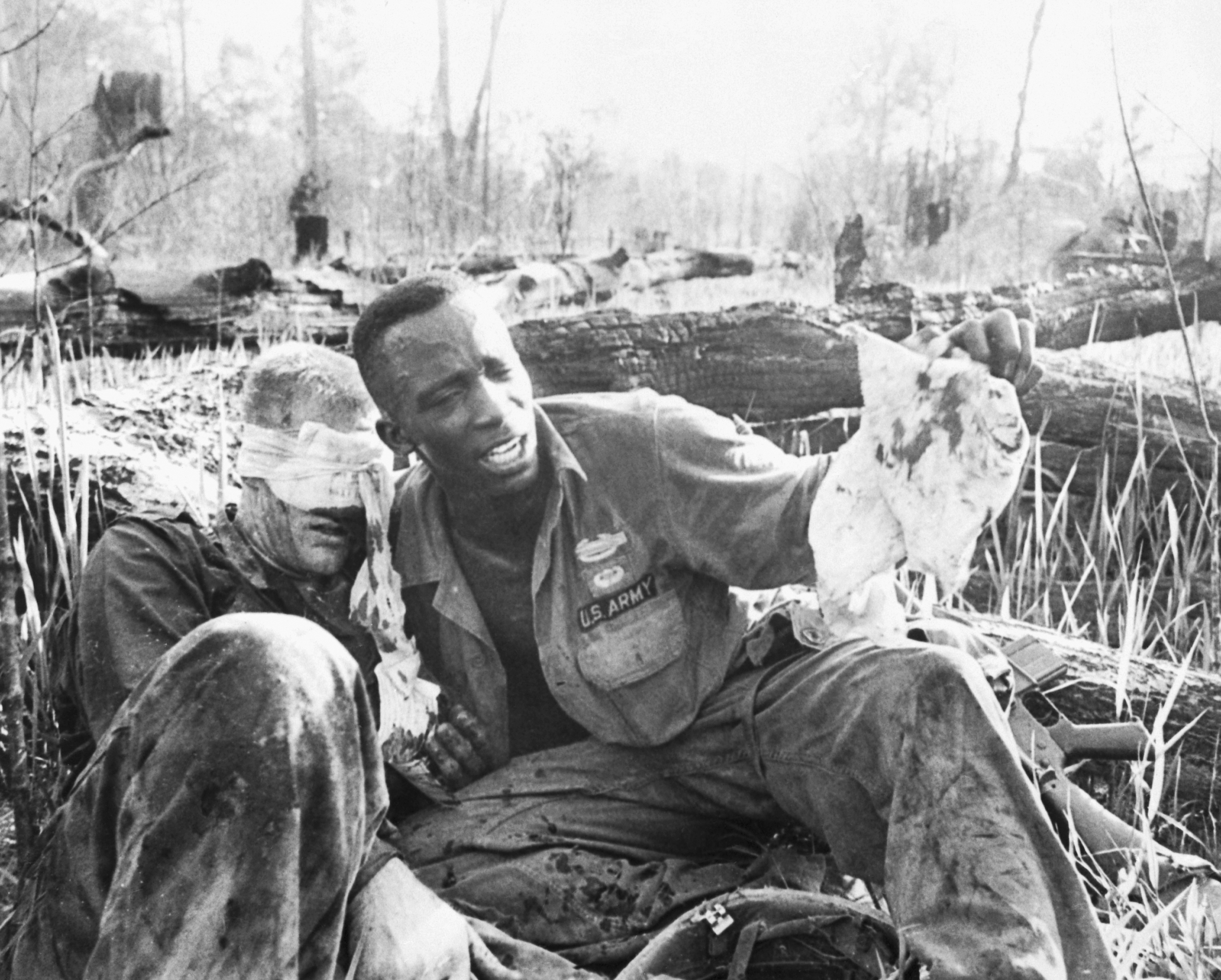 Black soldier sits with a wounded white soldier, who has a bandage over his eyes.