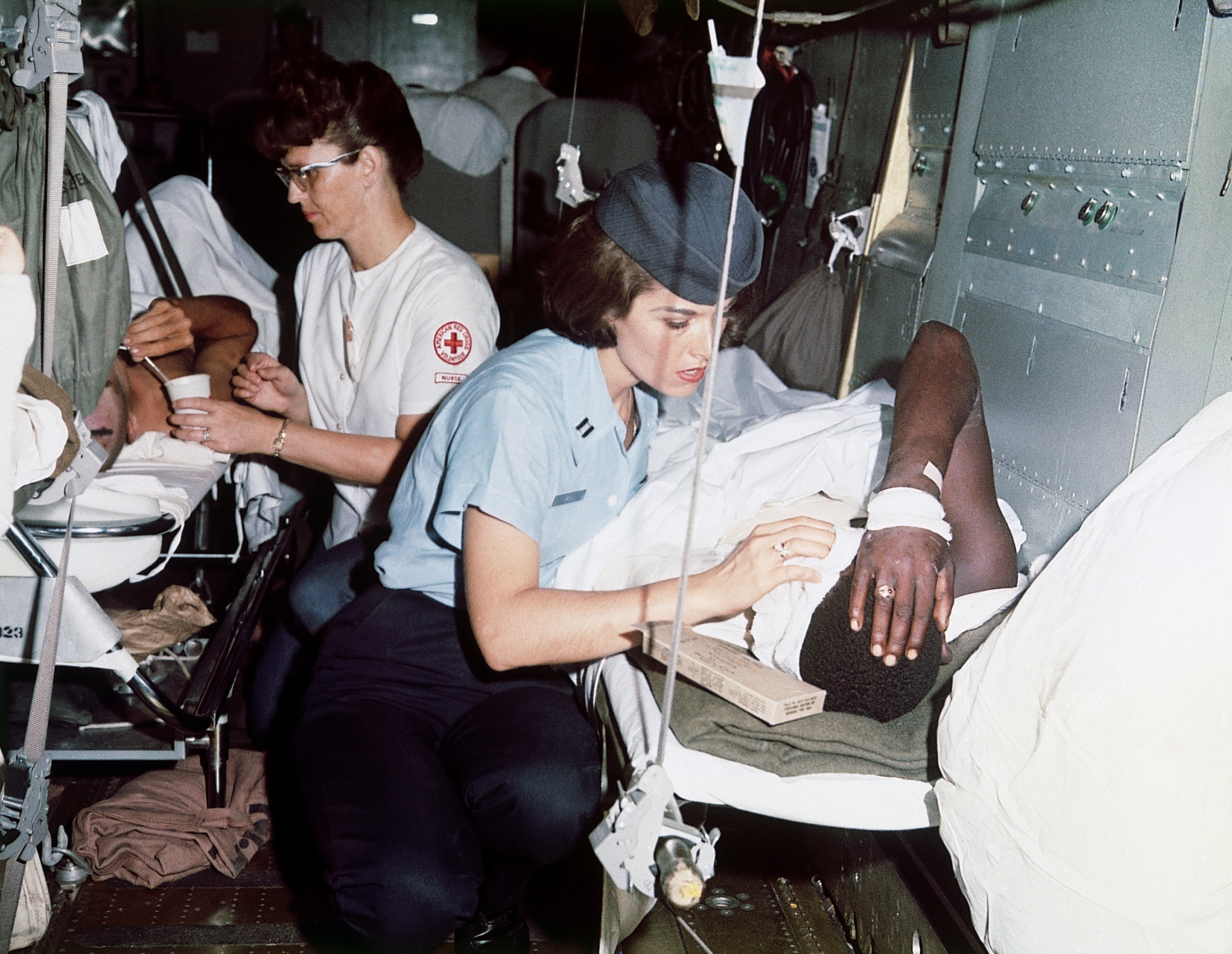 Nurses crouch beside wounded soldiers on gurneys in a transport vehicle.