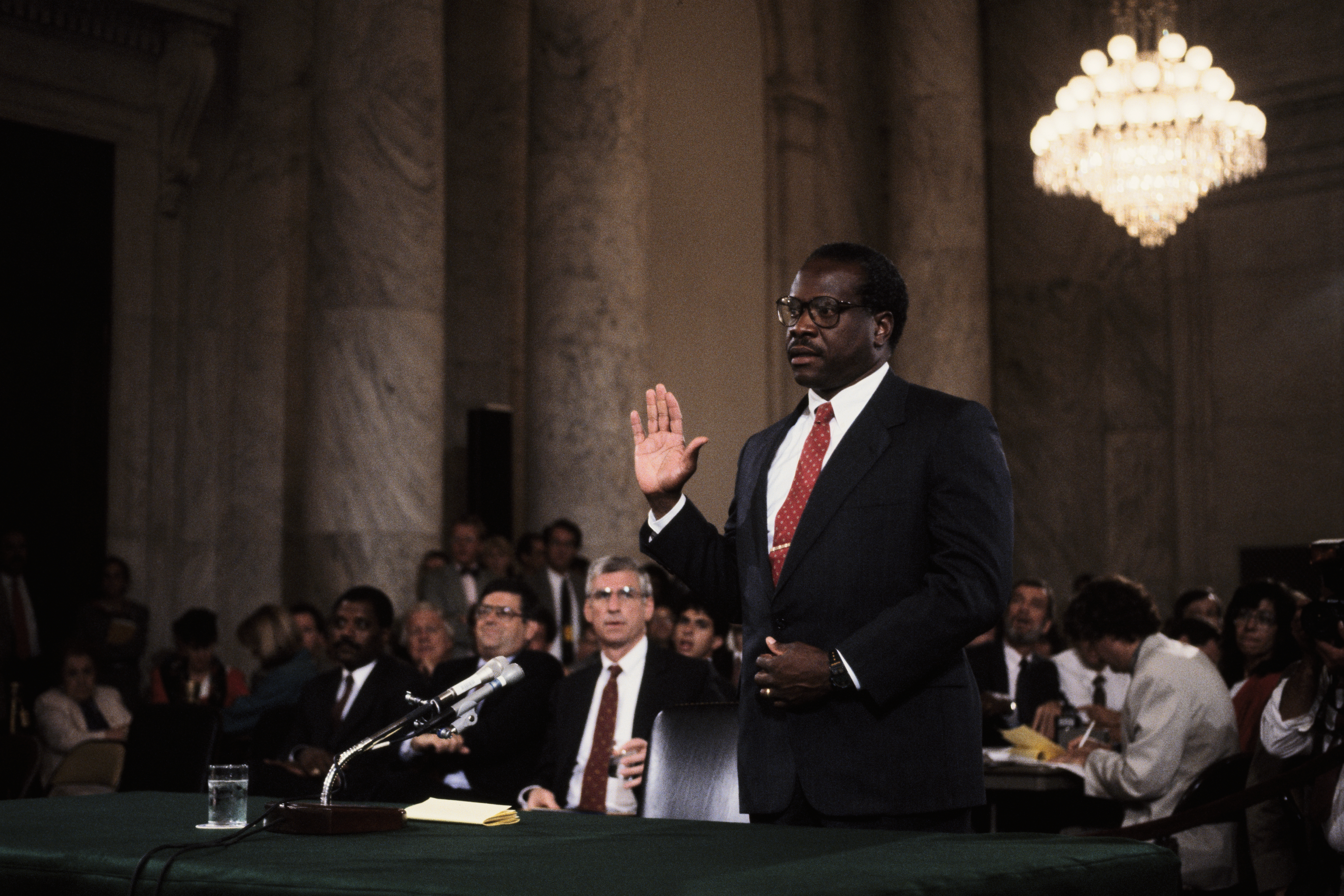 Supreme Court nominee Clarence Thomas, an African American man in a dark suit and red tie, wearing black-framed glasses, raises his hand while taking an oath during a hearing.