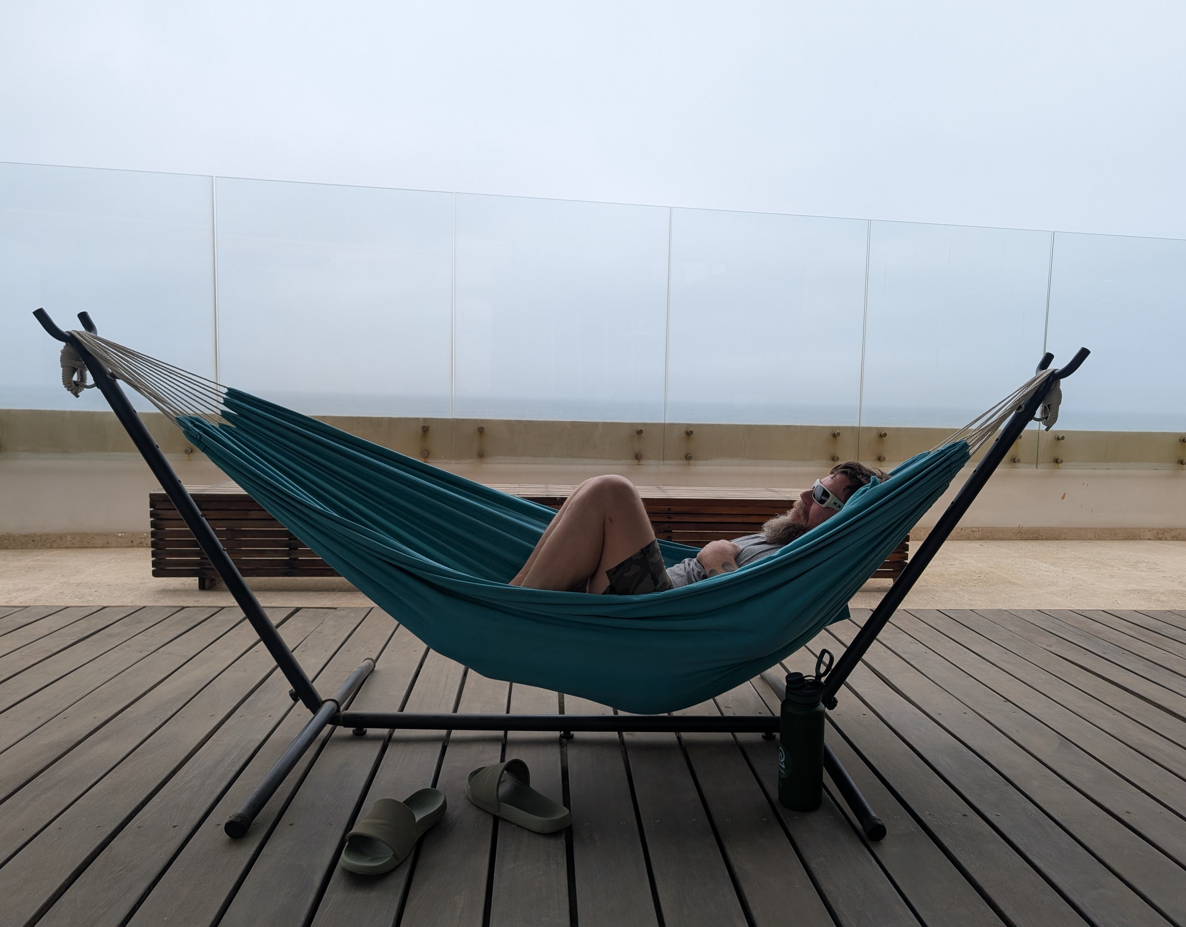 A bearded man in sunglasses relaxes in a hammock on a wooden deck with a view of the ocean.
