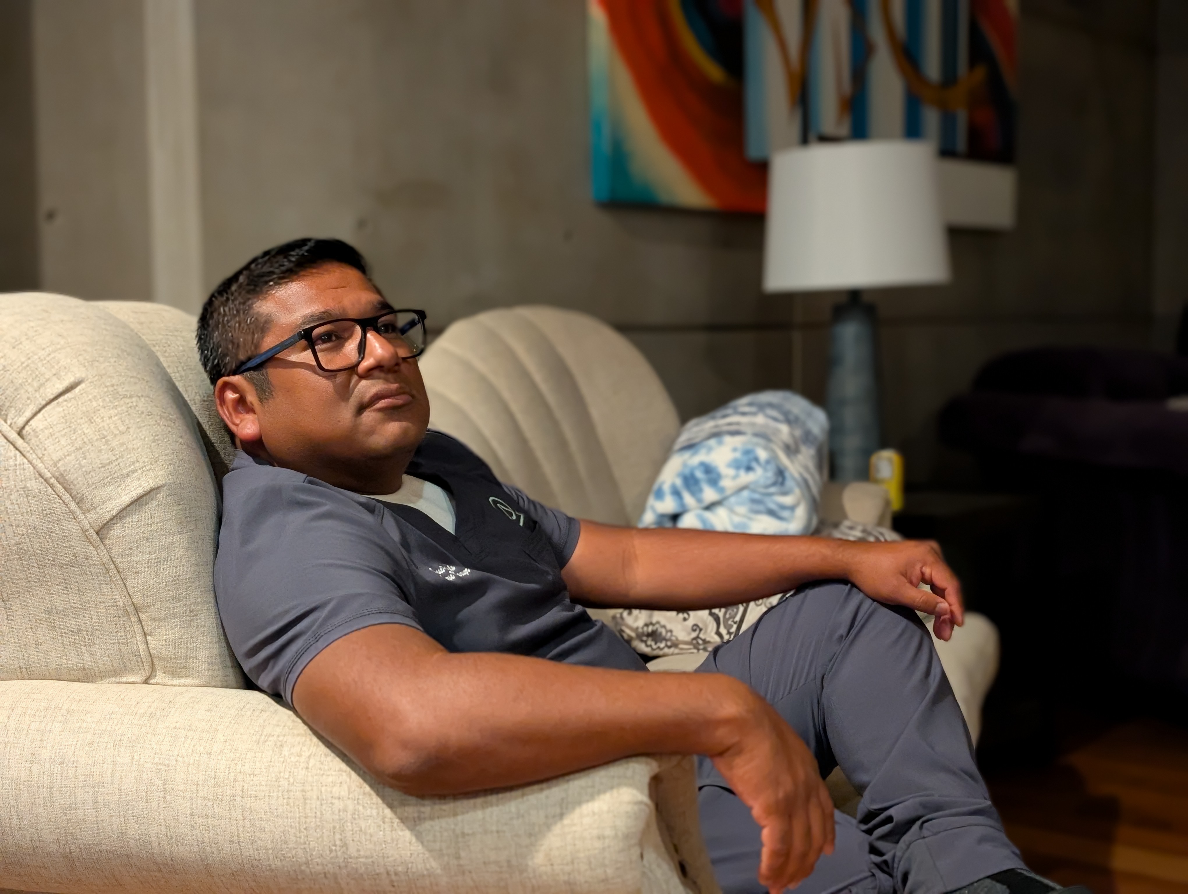 A bespectacled male nurse sits on a couch, looking off into the distance.