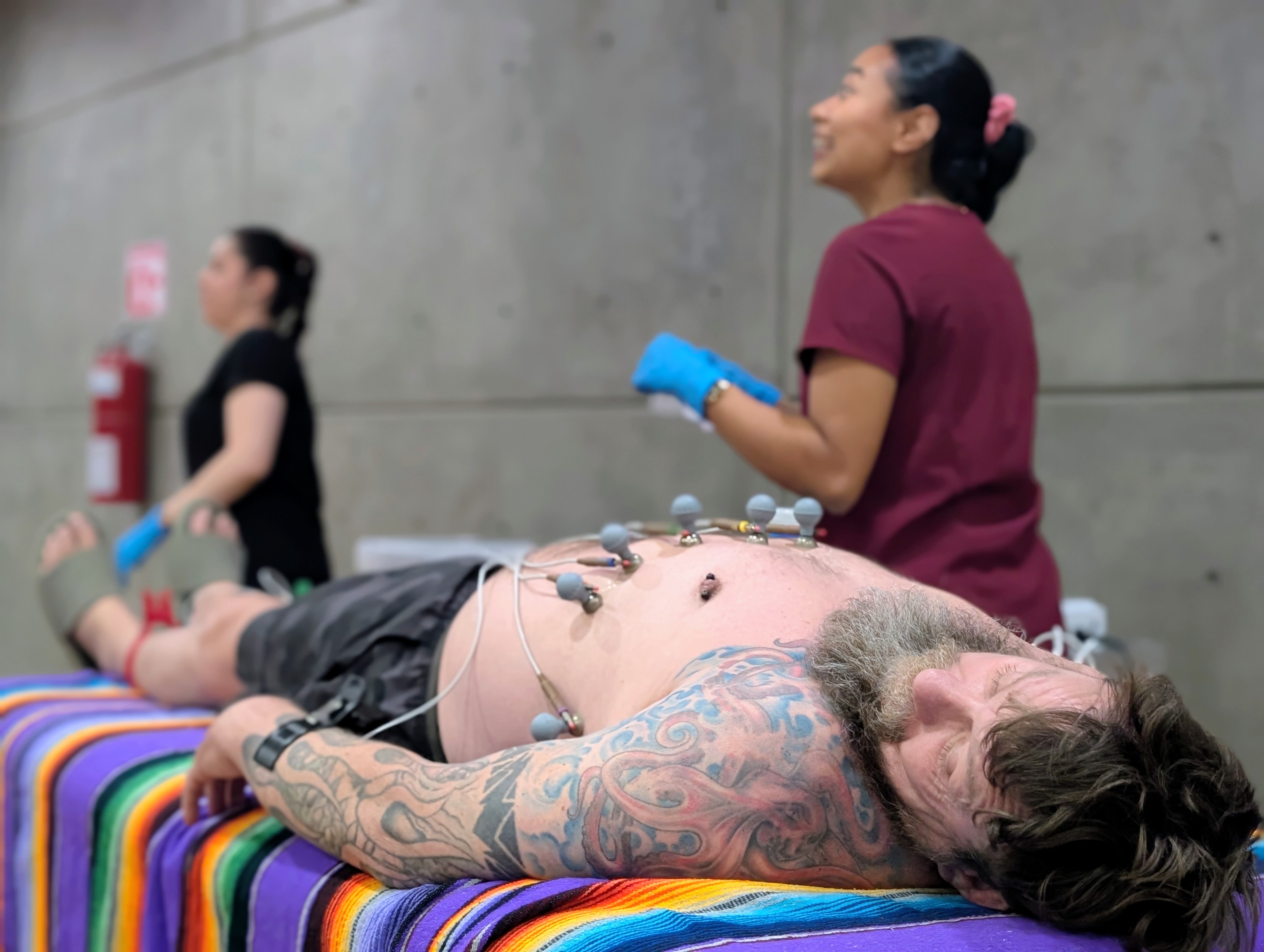 A bearded man lies on a colored blanket and is hooked up to an EKG.