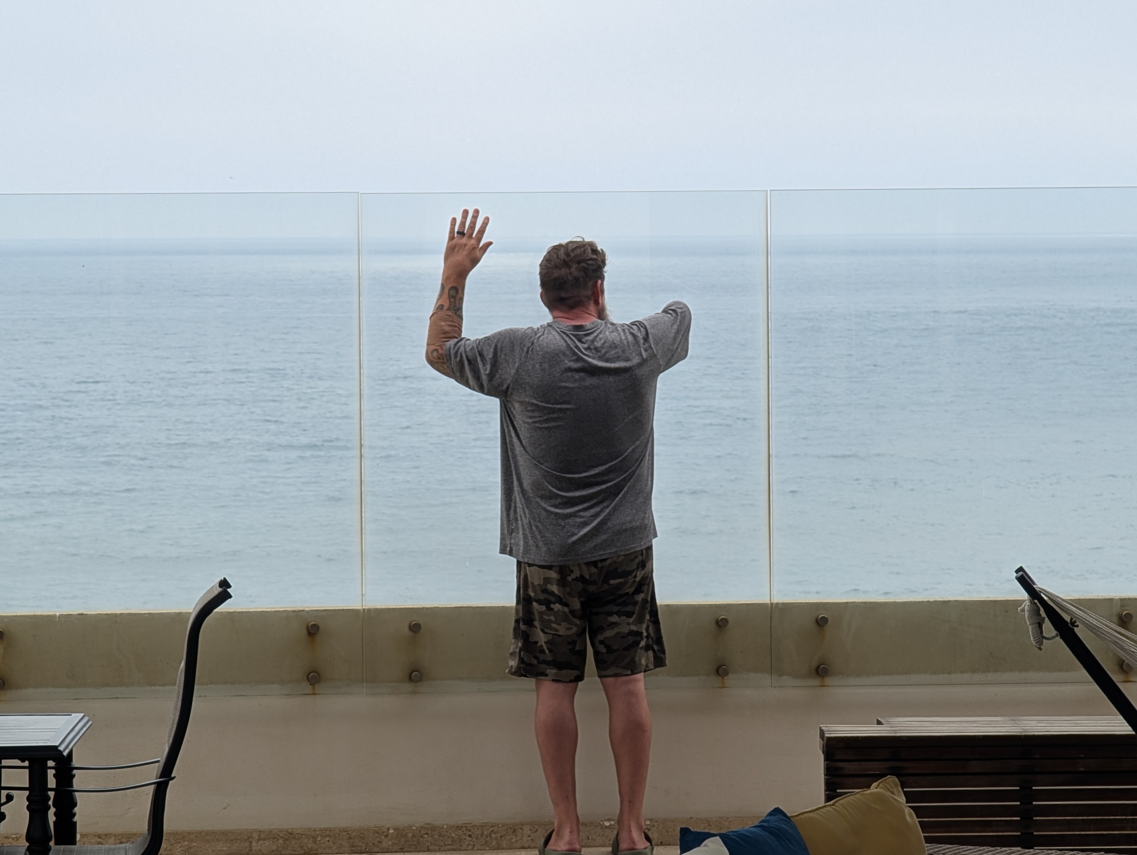 A man, missing his right arm, leaning against a glass wall, facing the ocean, looked out toward the waves.
