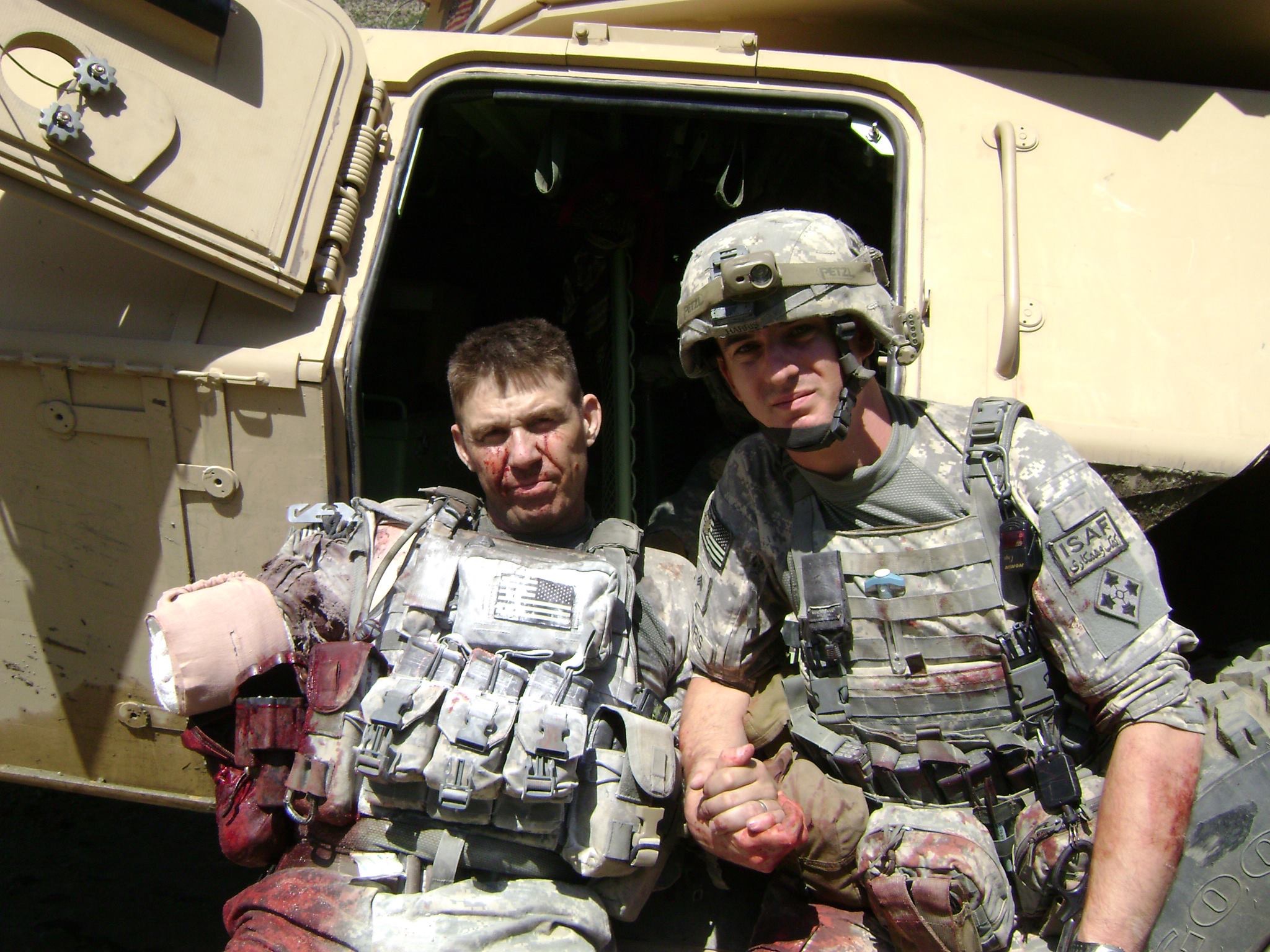 Two American soldiers in Army fatigues and gear lean against a Humvee. The man on the left is missing an arm, which is bloodied and bandaged, and holds the hand of the other man on the right.