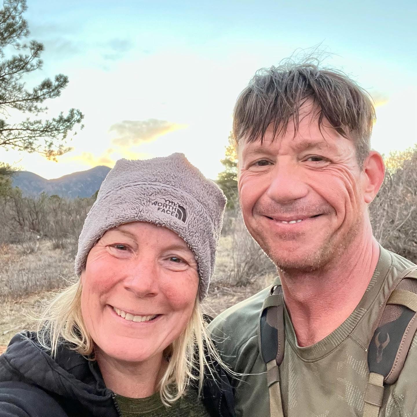 A selfie of two people on a trail in the mountains, a man and a woman, smiling at the camera.