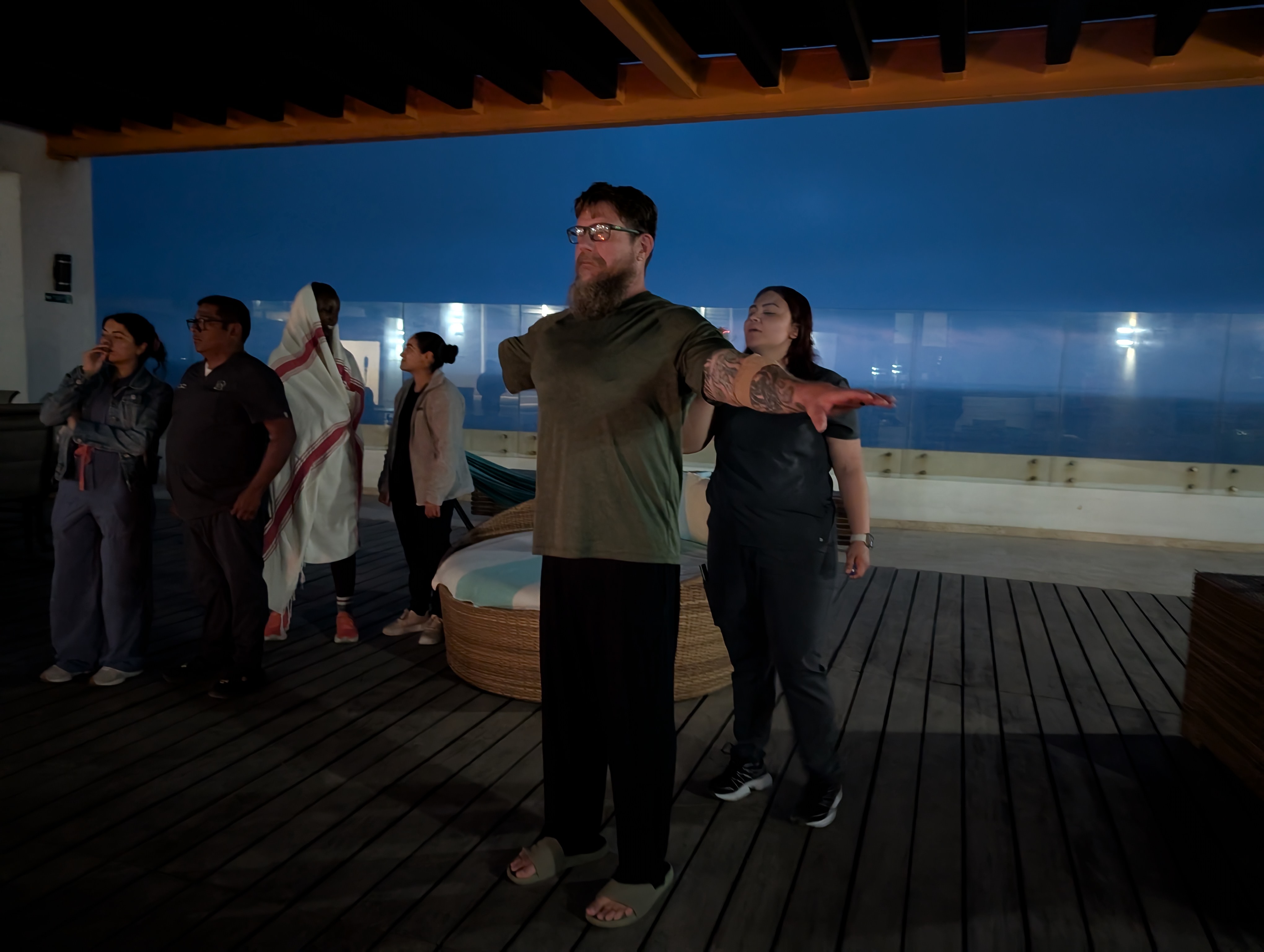 A bearded bespectacled man who is missing his right arm, stands with his limbs outstretched on a seaside deck while a nurse attends to him.