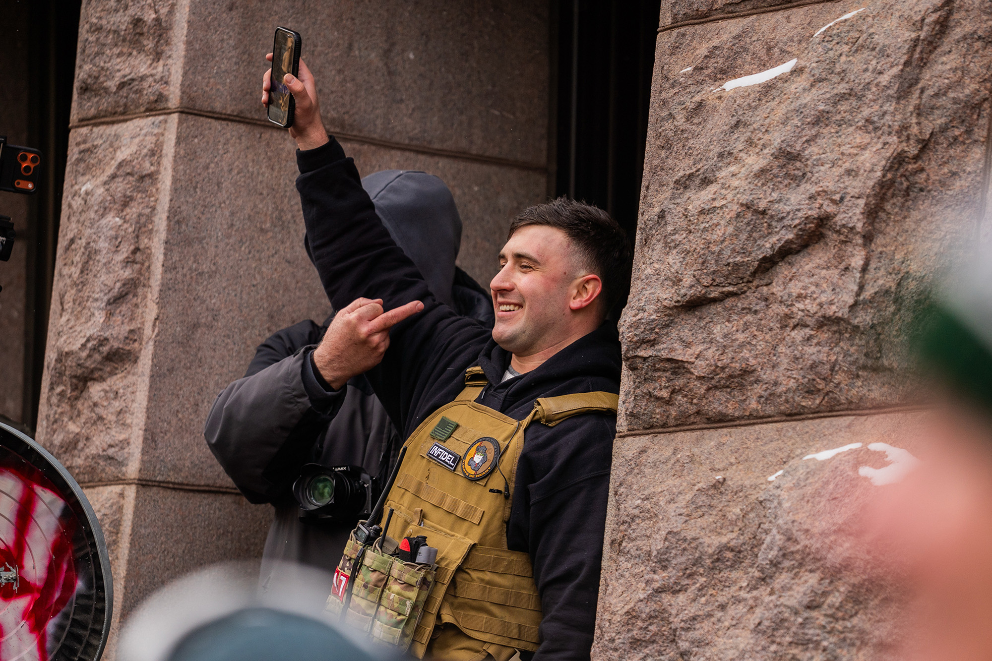 A man wearing a military vest smiles with his arm raised while another man flips him off.