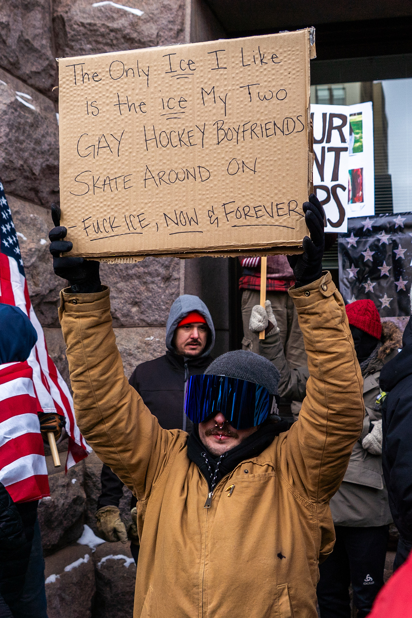 A person with ski goggles holds a sign that reads, "The Only ICE I like is the ice my two gay hockey boyfriends skate around on – Fuck ICE, now & forever."