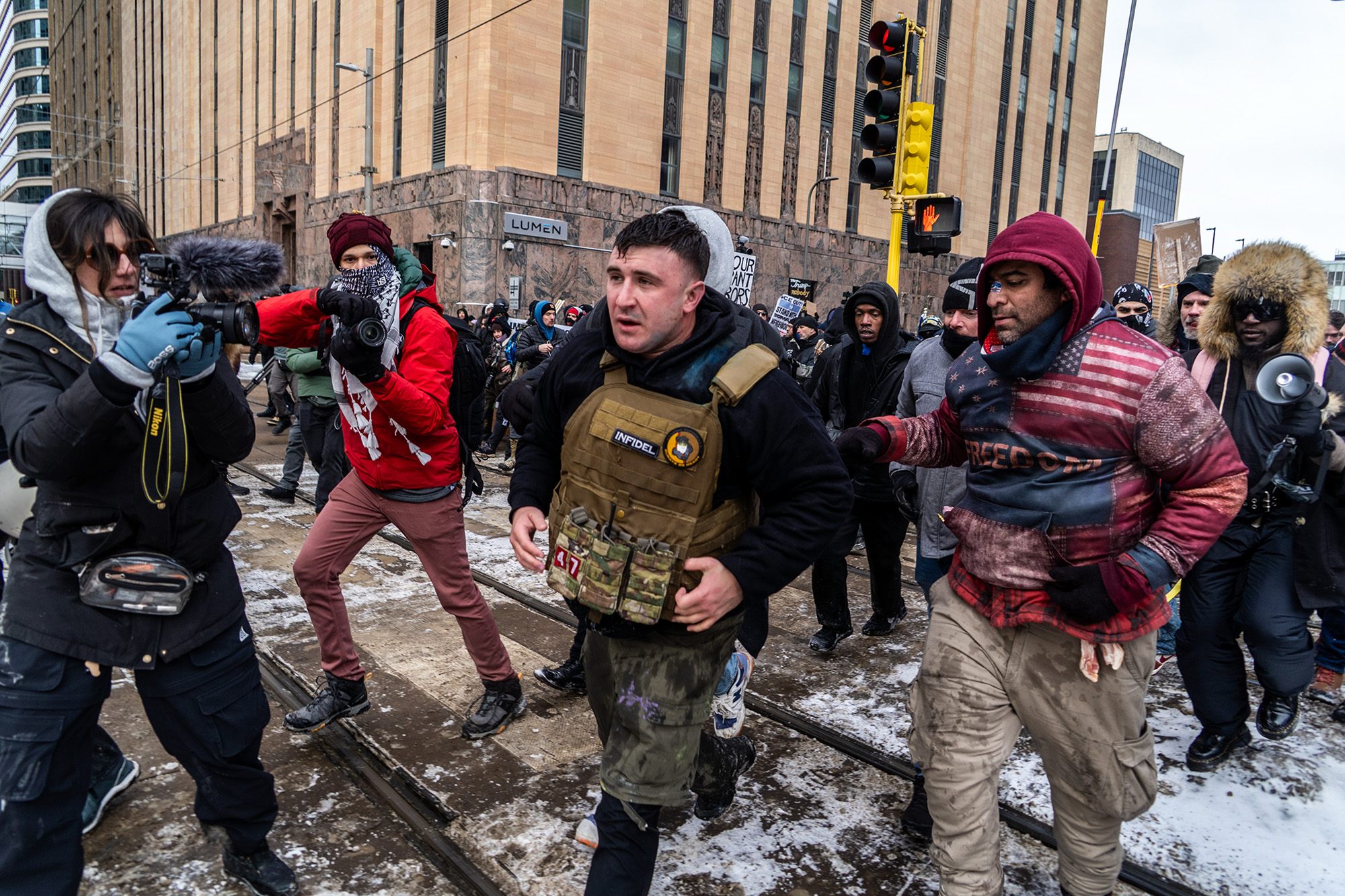 A man wearing a military style vest walks through snow surrounded and followed by a number of people.
