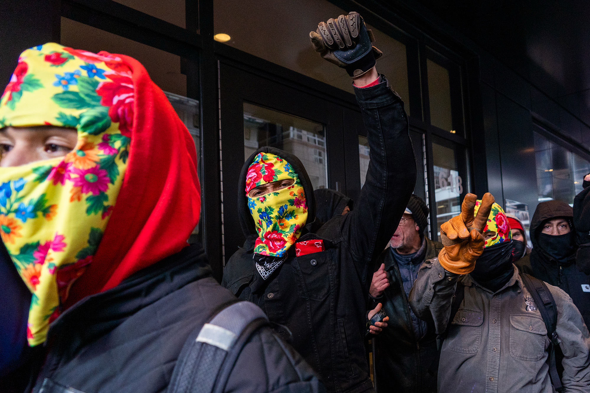 A group of three men wearing yellow floral masks walk with a raised fist and flashing a peace sign.