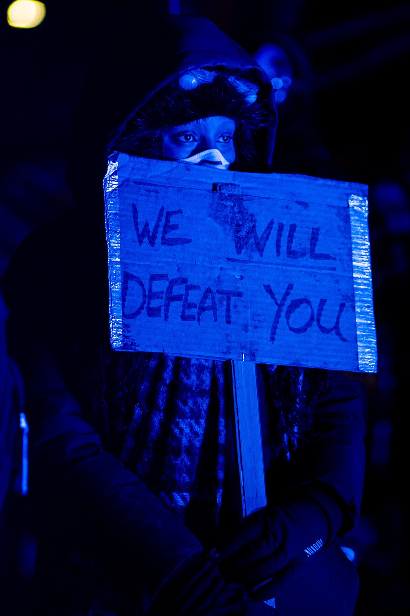 A woman holds a sign that reads, "We Will Defeat You" in the dark.