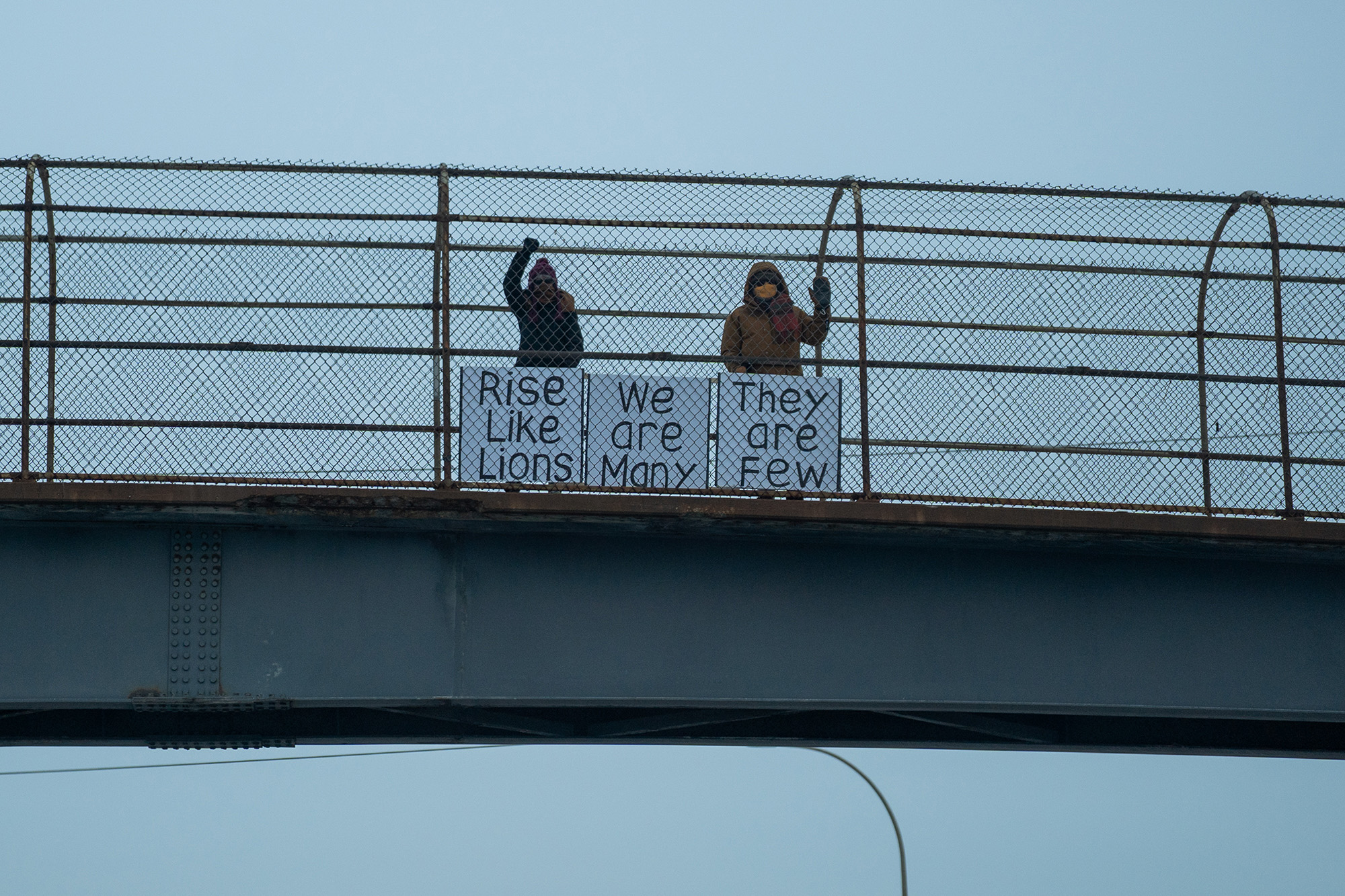 Two people on a high pedestrian overpass with signs that read, "Rise Like Lions," "We are Many," "They are few."