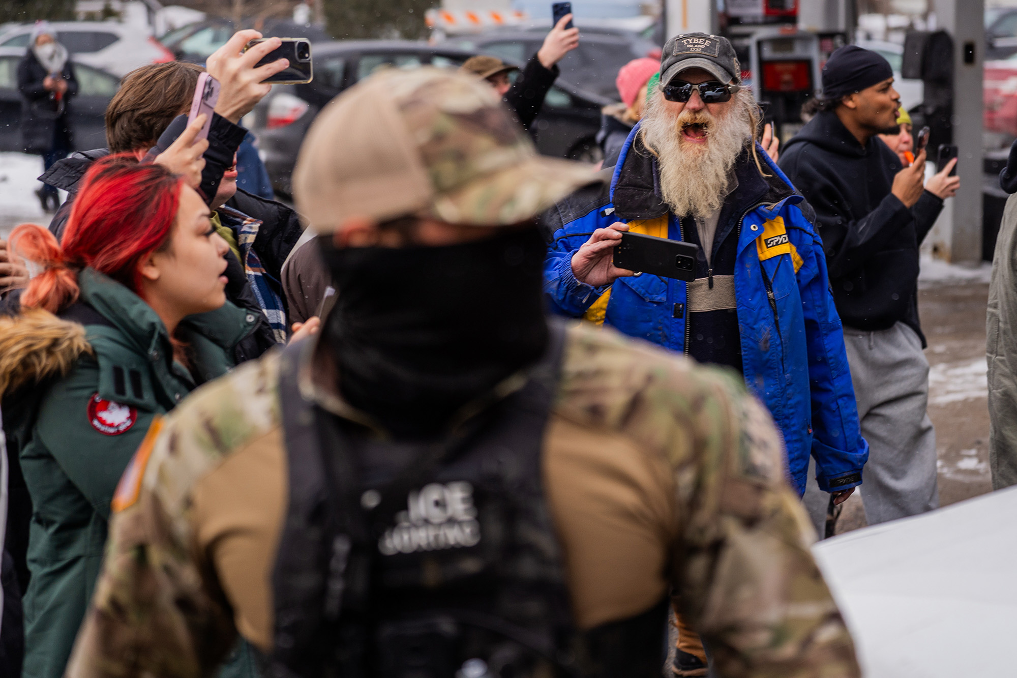 A man with a long beard wearing a blue jacket, holding a cellphone, yells at an ICE agent.