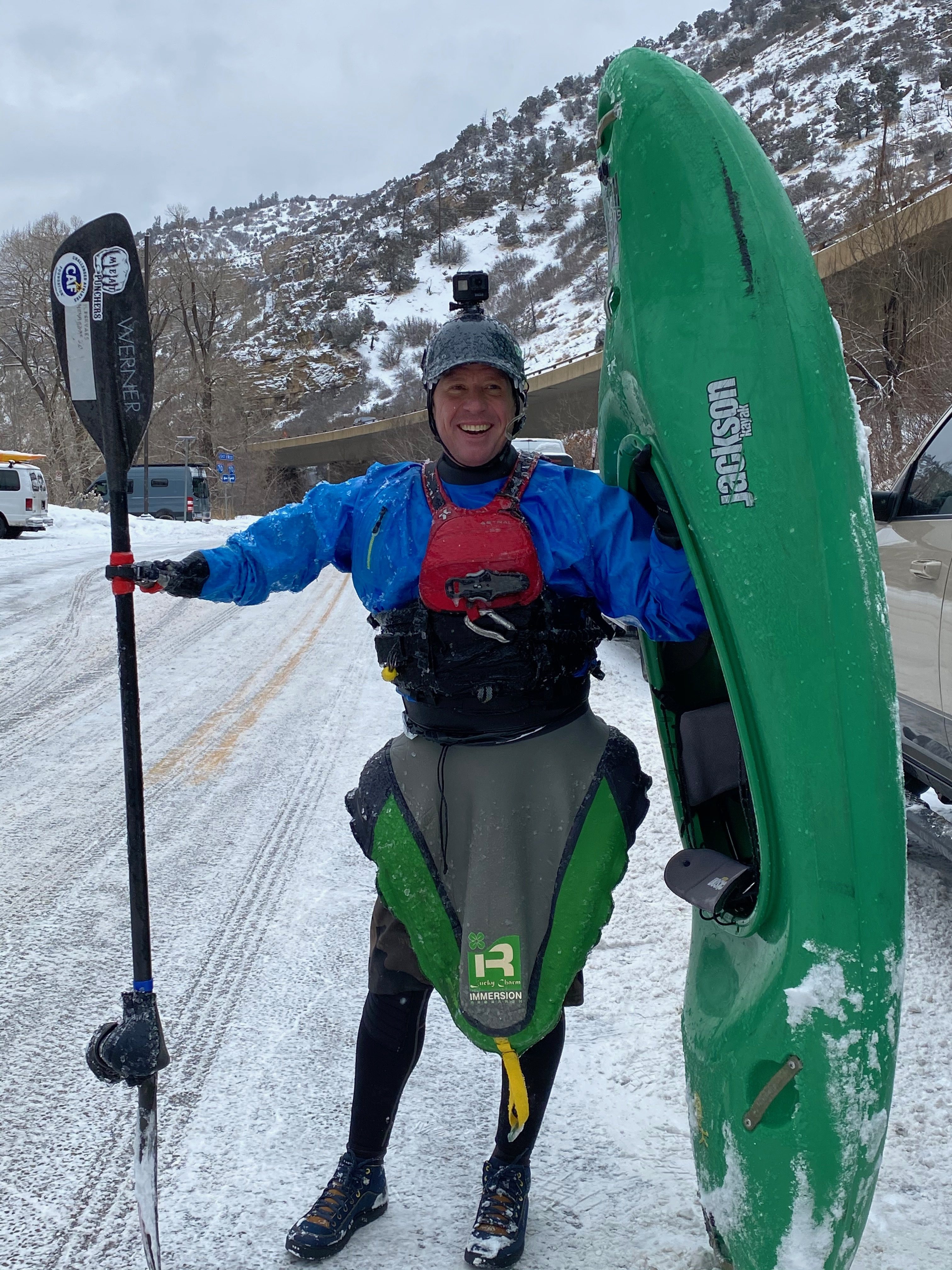 A disabled kayaker stands with his kayak in one hand and modified paddle attached to the other limb. There is snow on the ground and he is dressed in winter gear and a helmet.