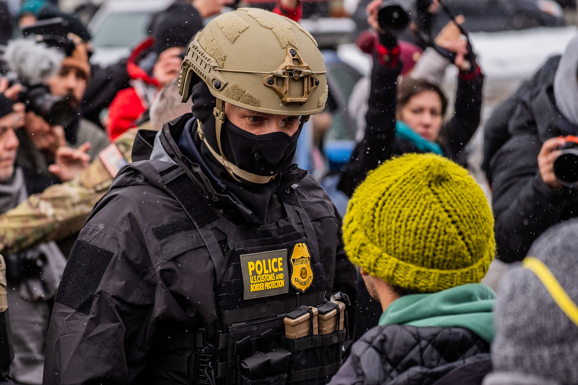 Masked federal agent looms over a protester in a yellow hat.