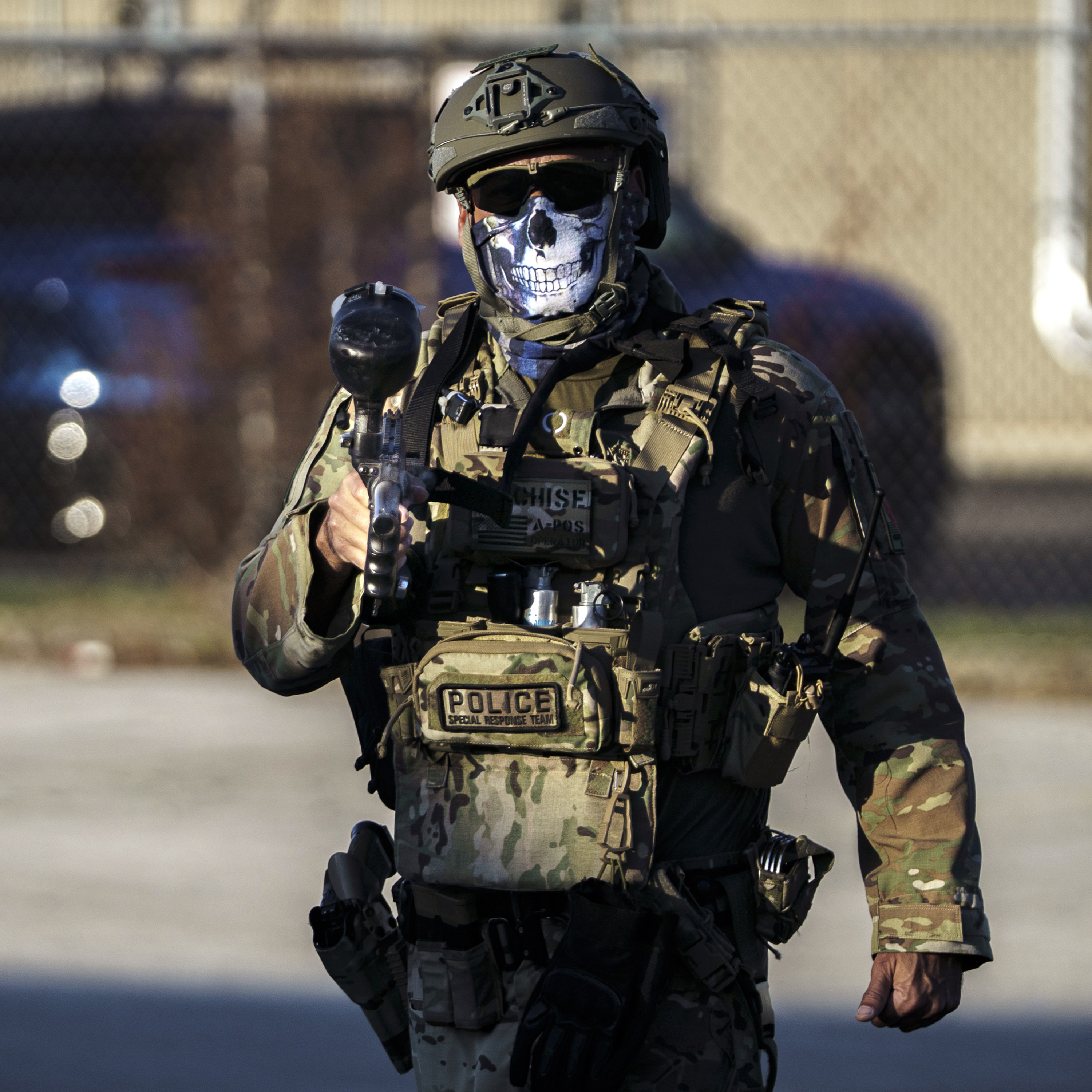 ICE agent in full camouflage with a helmet, sunglasses and a skull face mask, holding a pepper ball gun.