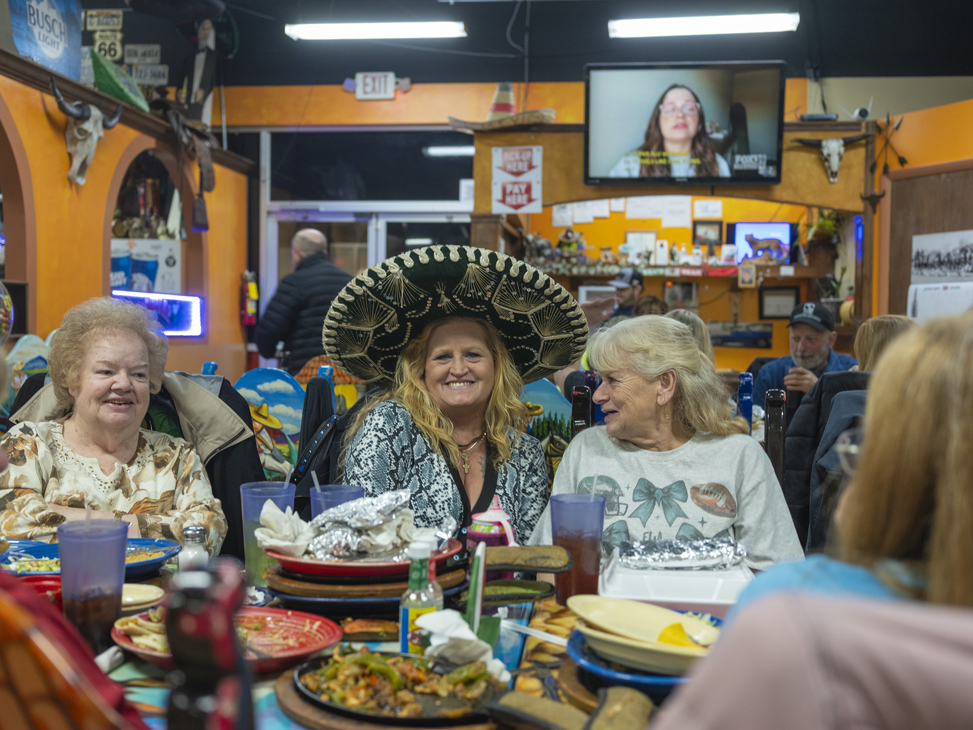 Three women smiling, sitting at a table in a Mexican restaurant. The woman in the center wears a black embroidered sombrero.