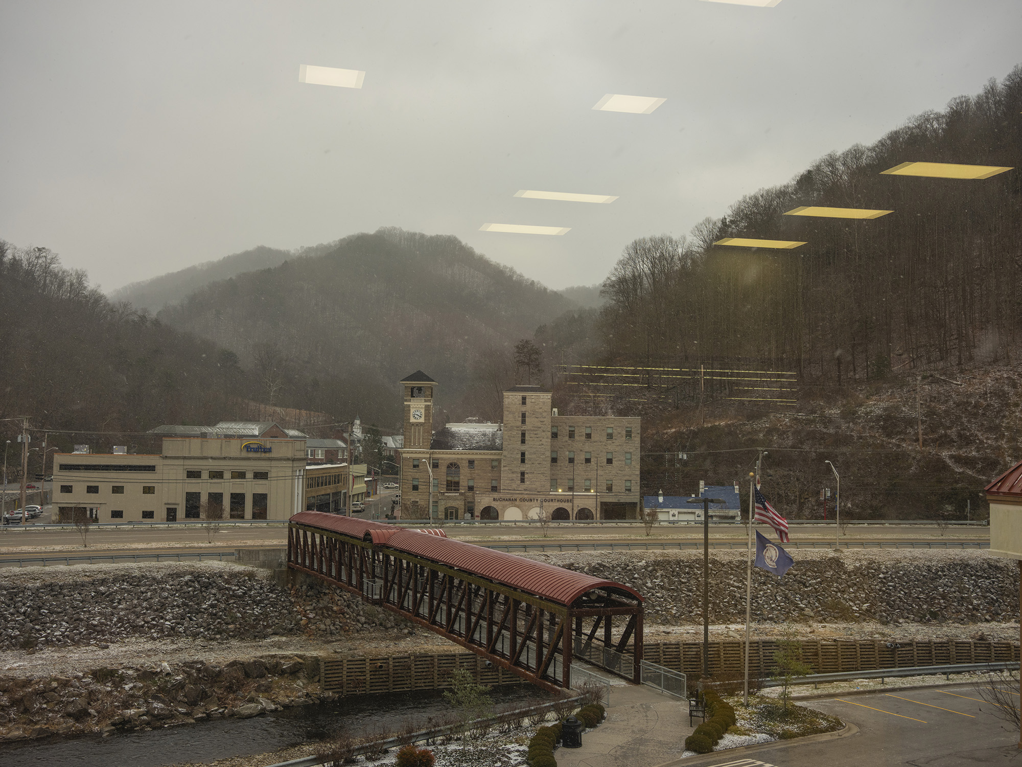 A small town nestled in the mountains alongside a river on an overcast day, seen through glass with reflections of overhead lights.