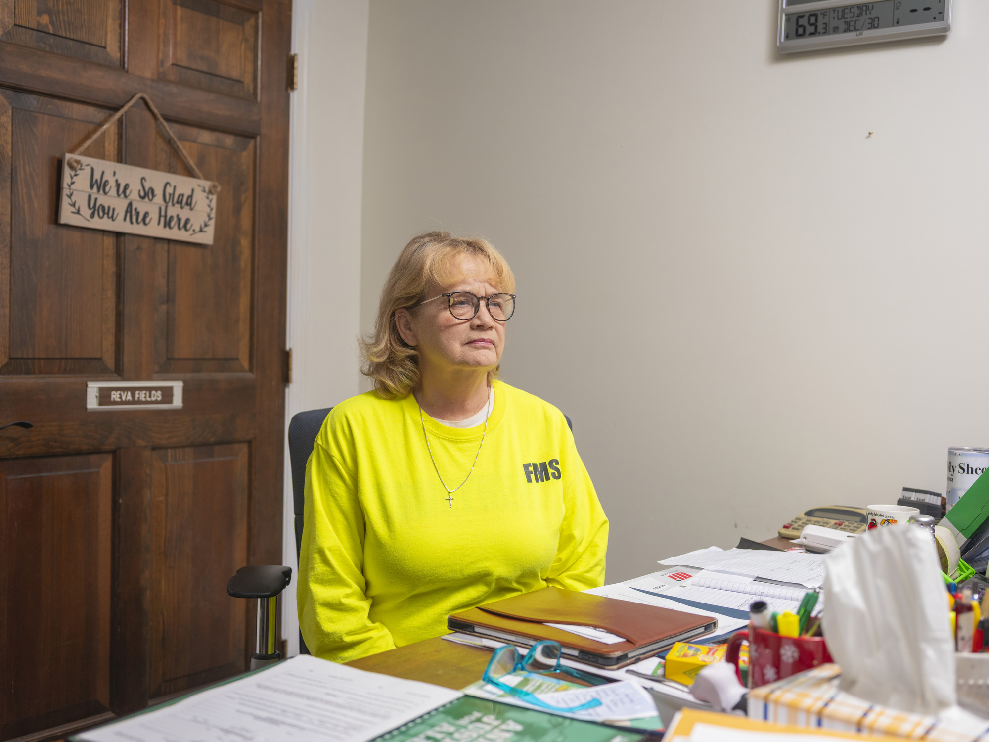 Portrait of a woman in a yellow sweatshirt sitting at a table with paperwork spread across it. A decorative sign hanging on a door behind her reads, "We're so glad you are here."