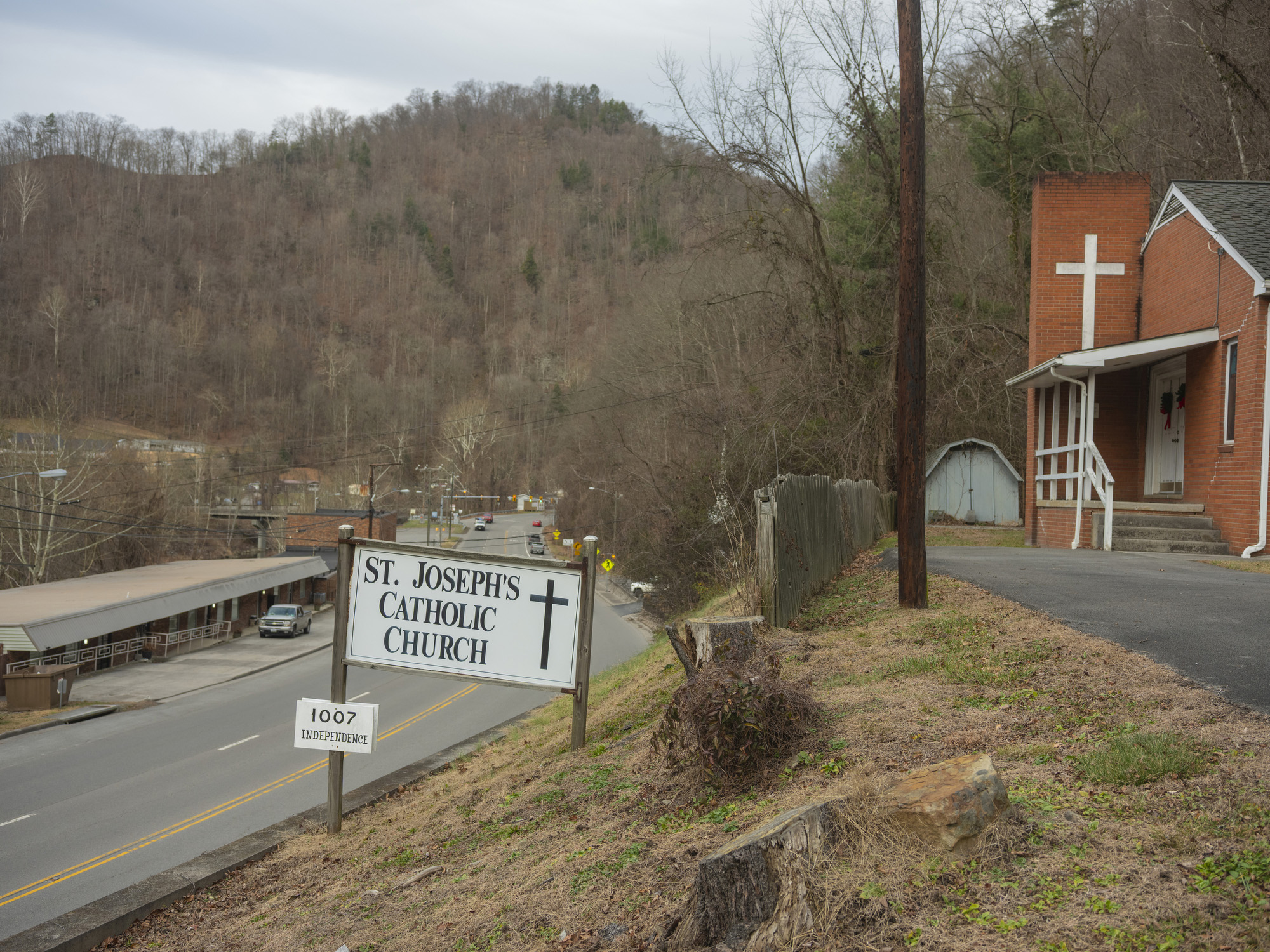 A red brick church on a hillside with a road running just below it. A sign out front reads, "St. Joseph's Catholic Church."